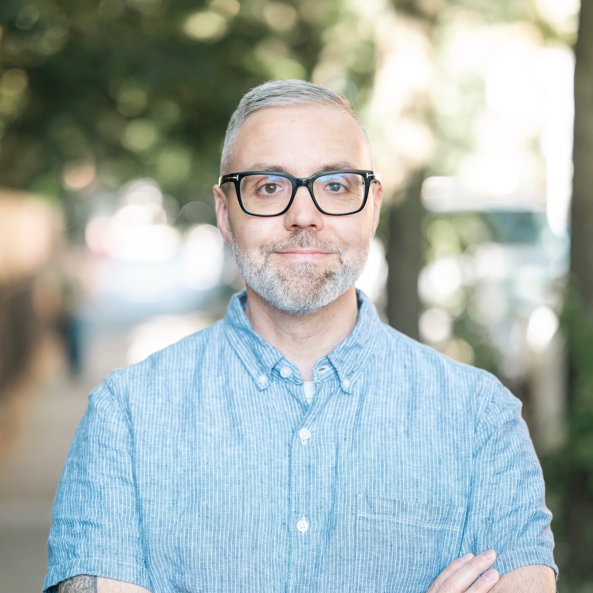 Man with glasses and gray hair, wearing a blue shirt, standing outdoors with blurred trees and sunlight in background.
