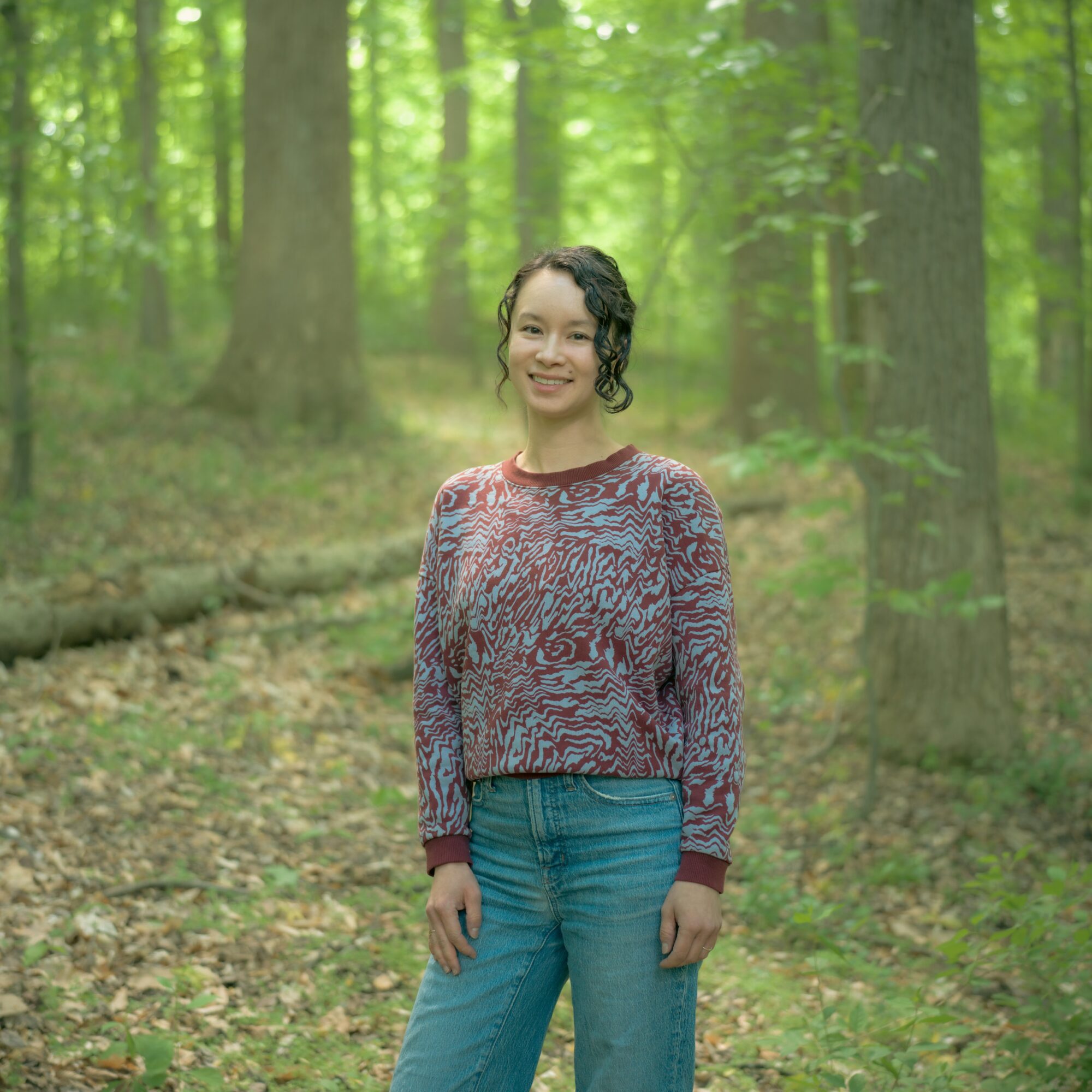 Young woman standing in a green forest, smiling, wearing a patterned sweater and jeans.
