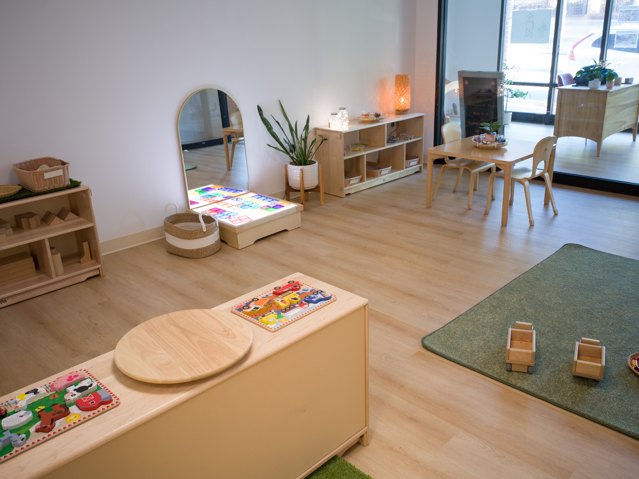 Children's playroom with furniture, books, plants, and a green rug, near a window with outdoor view.