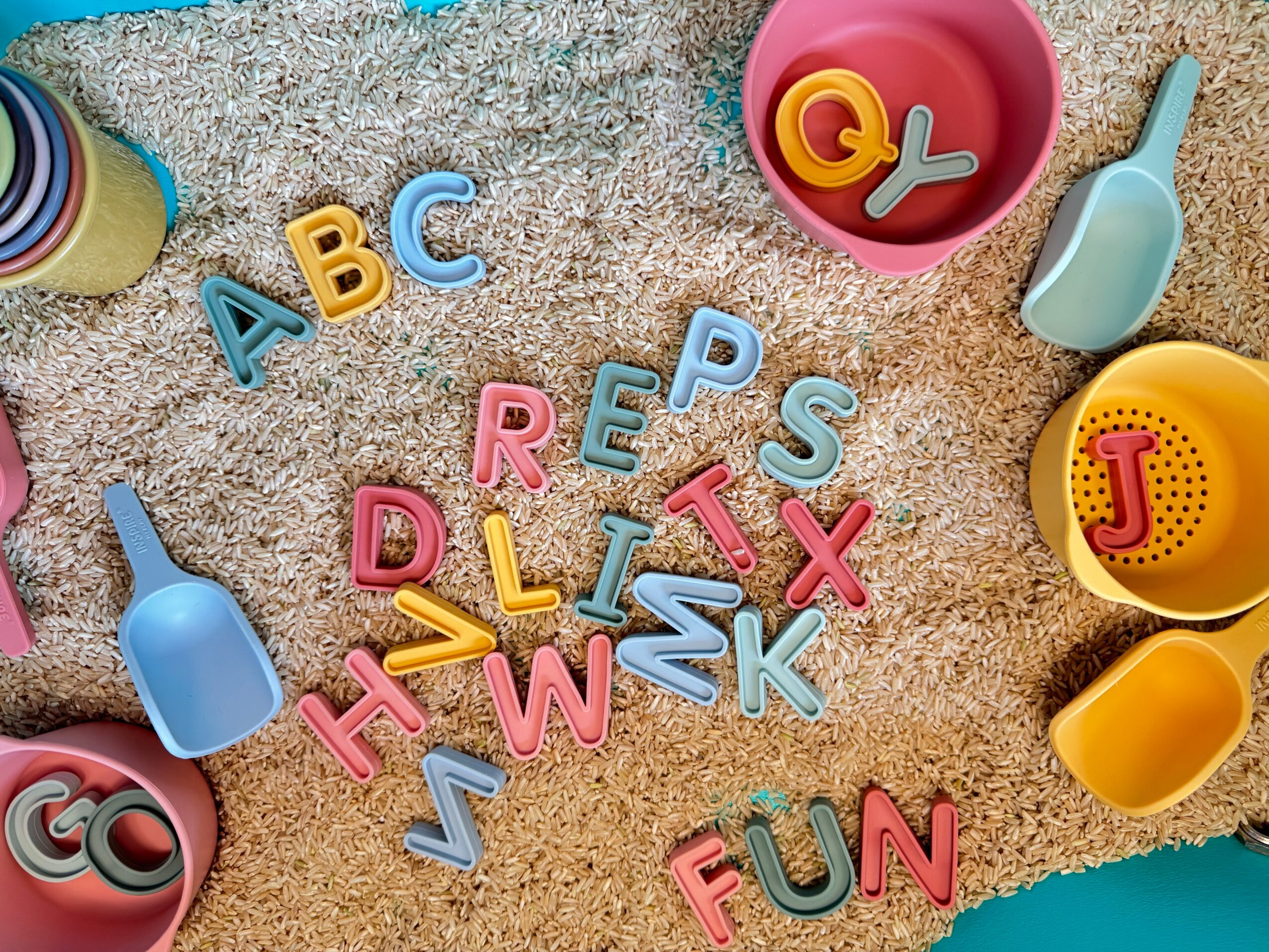 Colorful plastic buckets, shovels, and alphabet letters scattered on a sandy surface.