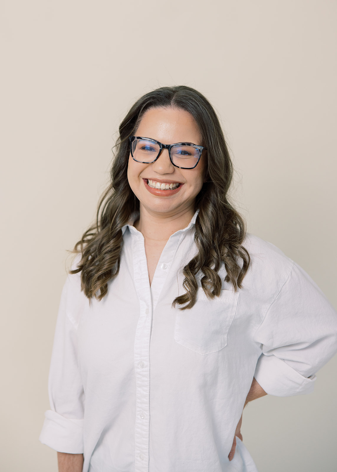Smiling woman with glasses and wavy hair wearing a white shirt, standing against a plain background.