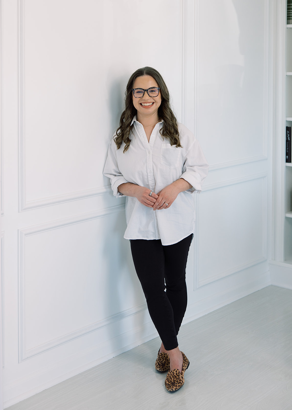 Woman with glasses and long hair, smiling, standing against a white wall in a bright room, wearing a white shirt, black pants, and leopard print shoes.