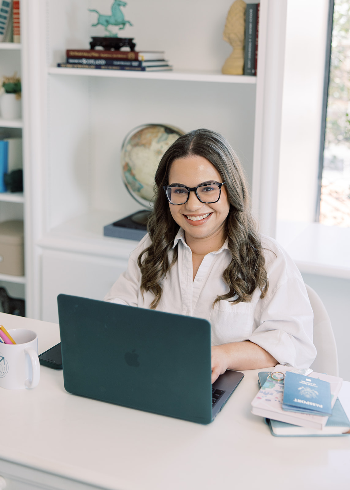 Young woman with glasses smiling at laptop in a bright room with books and globe, sitting at a white desk.