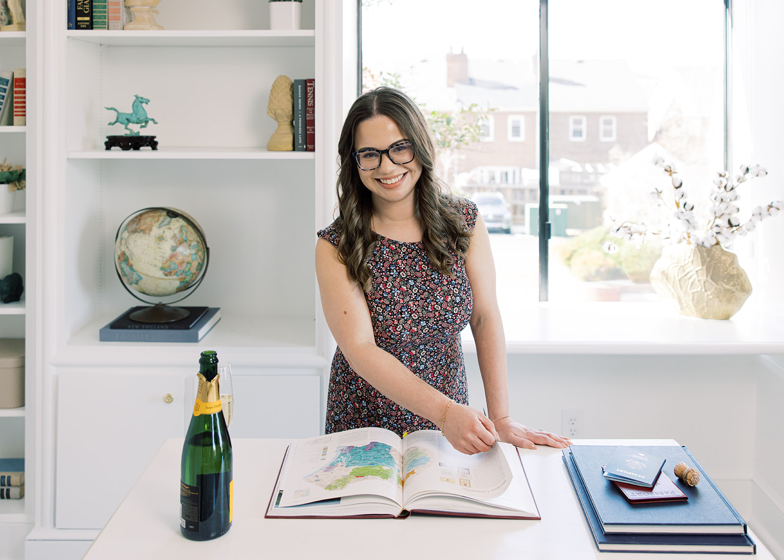 Smiling woman with glasses stands at a desk with an open book, a bottle, and a tablet, in a bright room.