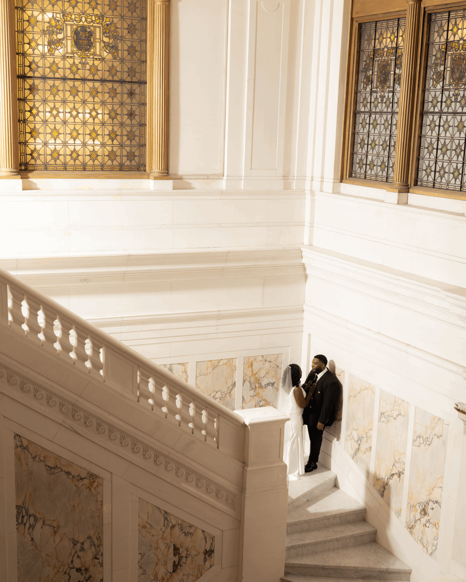 Two people stand on stairs near large windows with decorative glass, inside a bright, ornate interior with white walls and marble accents.