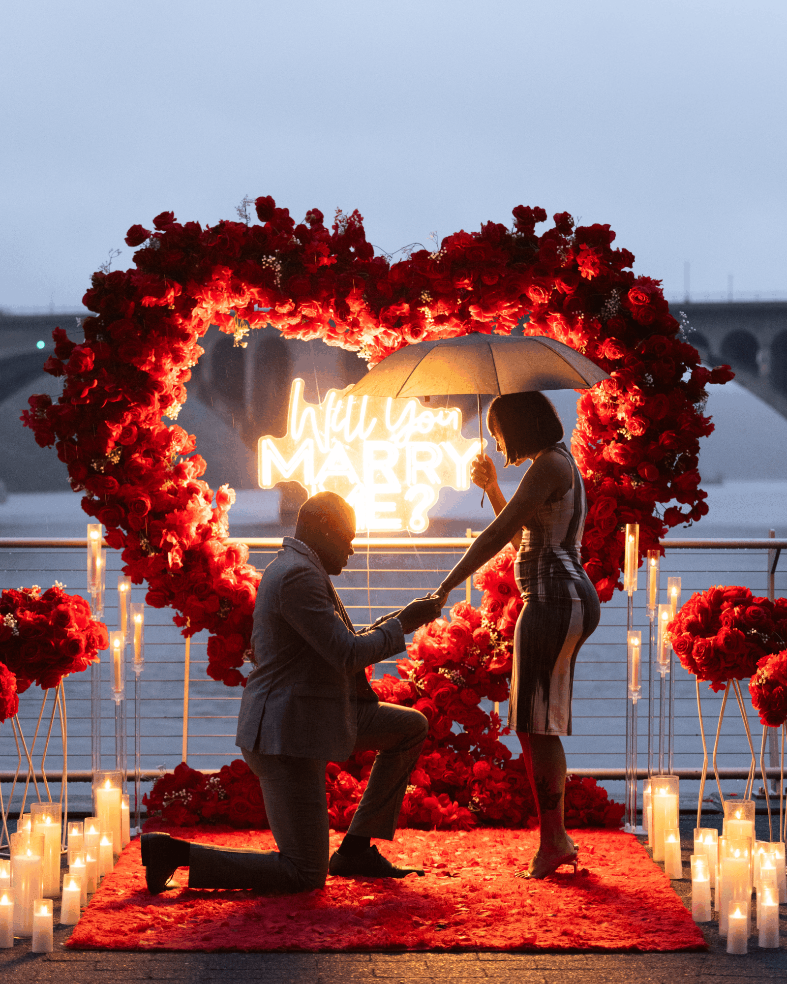 Person proposing to another person under a heart-shaped floral arch with candles, on a red carpet near water, evening setting.