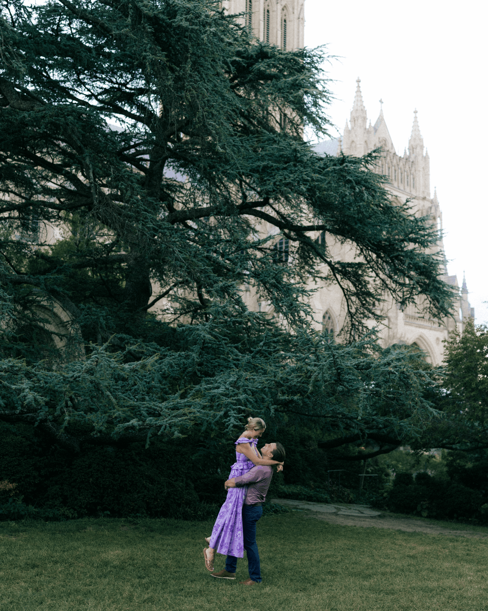 Two people embrace on a grassy area with large trees and a castle-like building in the background.