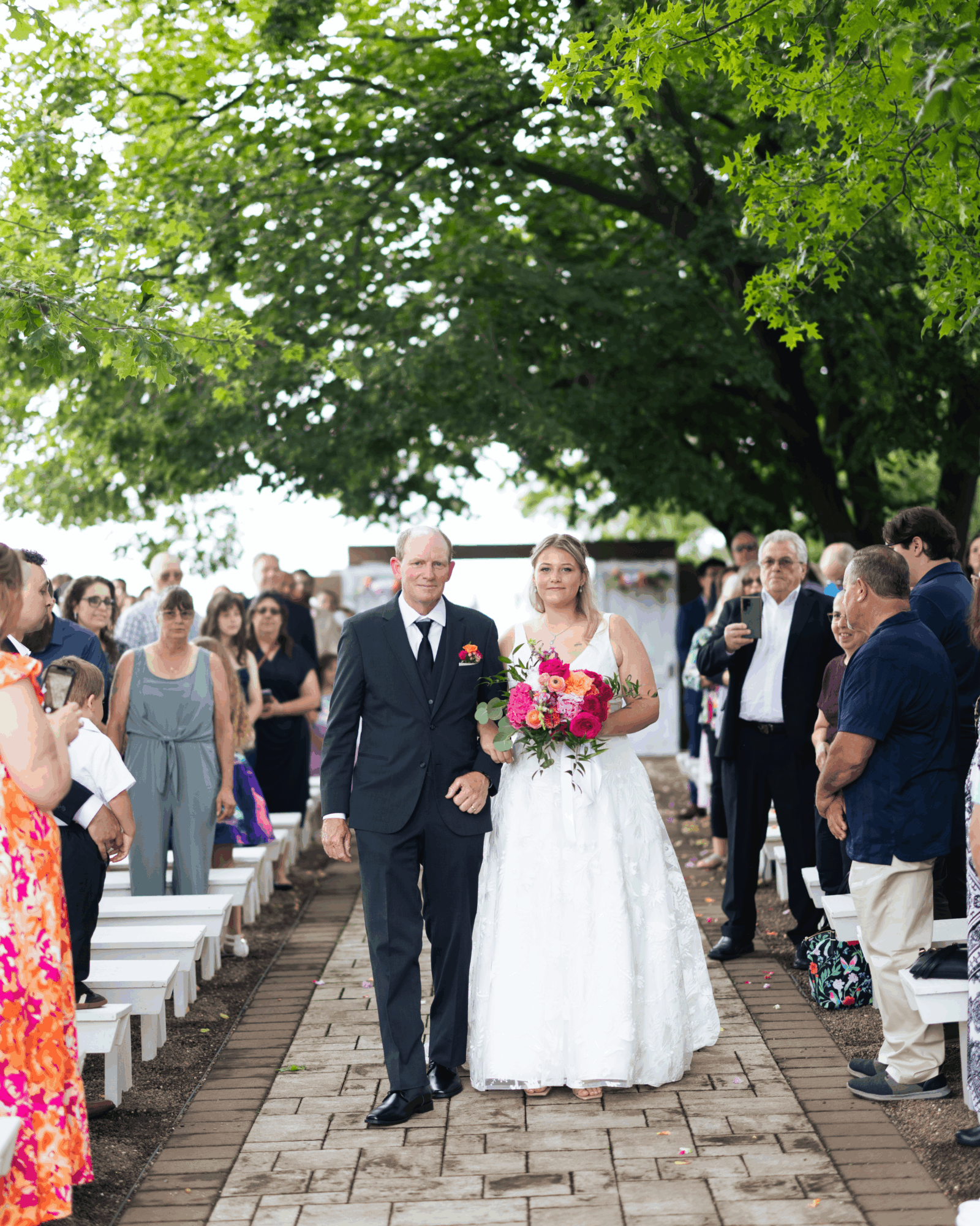 Bride in white dress holding a bouquet walking with a man in a suit, surrounded by guests outdoors under a large tree.