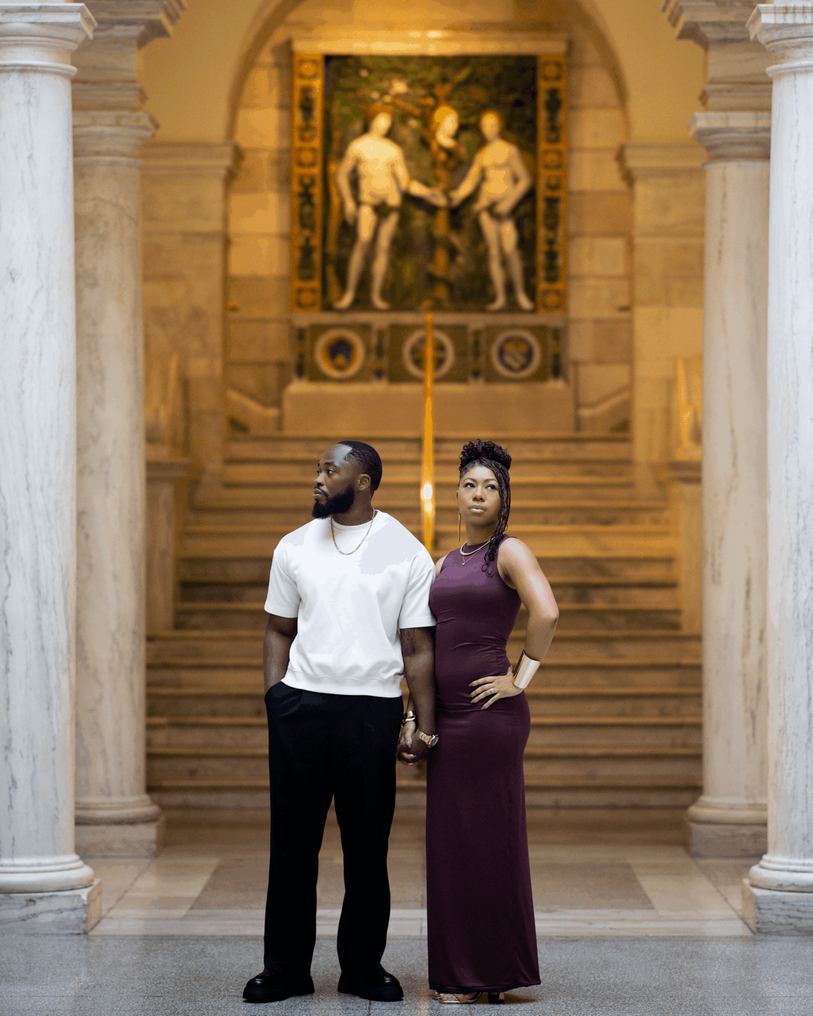 A man and woman stand in a grand hall with marble columns and a staircase, holding hands, with a painting in the background.