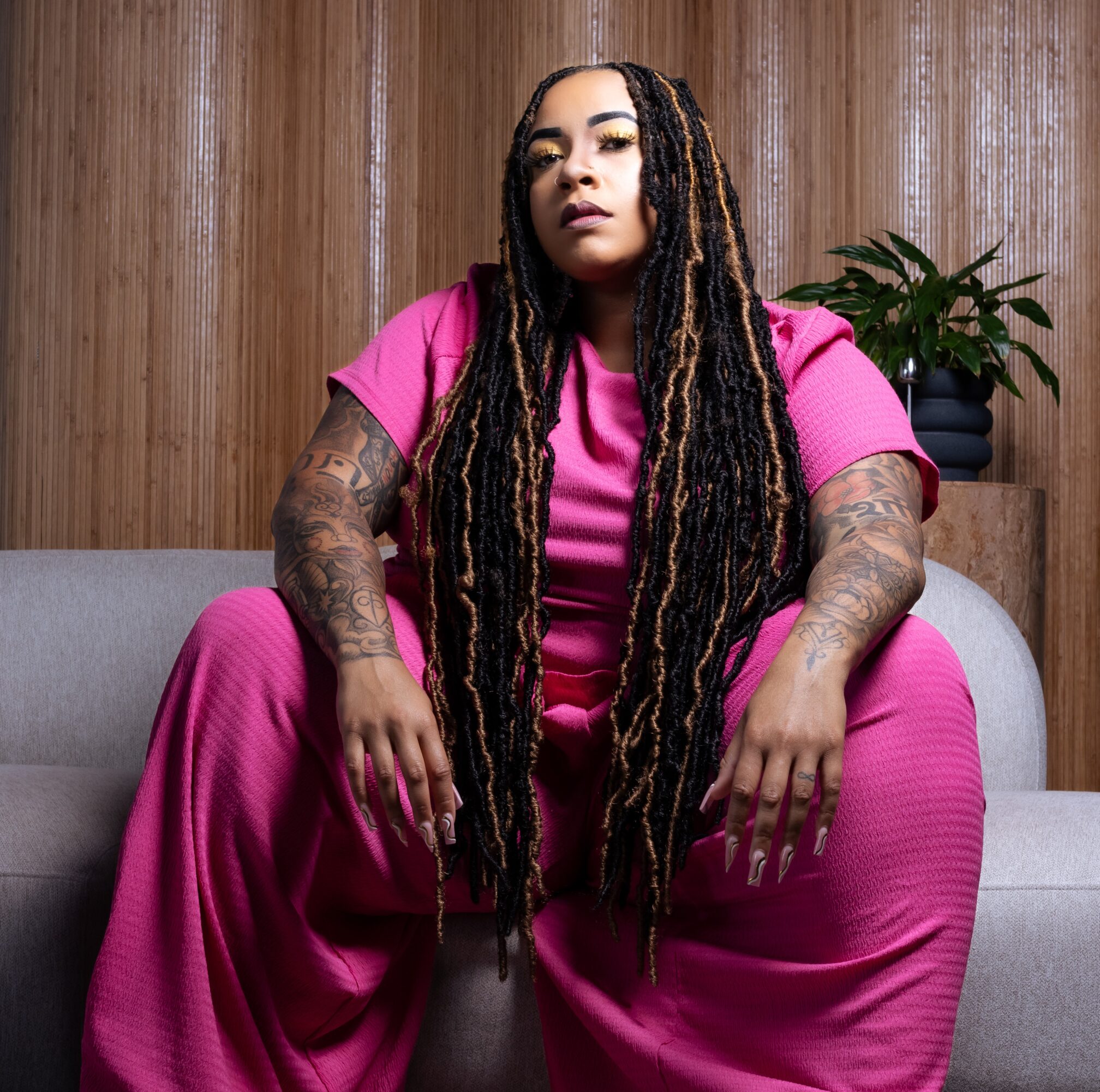 Woman with long dreadlocks wearing a pink outfit, sitting on a sofa with a wooden wall and plant behind her.