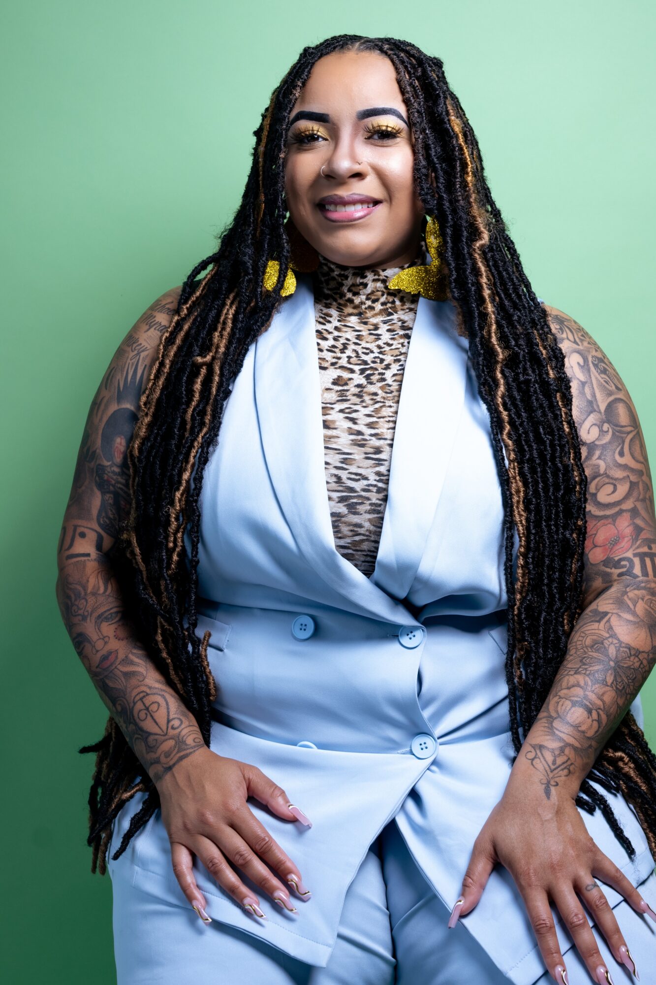 Woman with long dreadlocks, wearing a light blue blazer over a leopard print top, smiling, against a green background.
