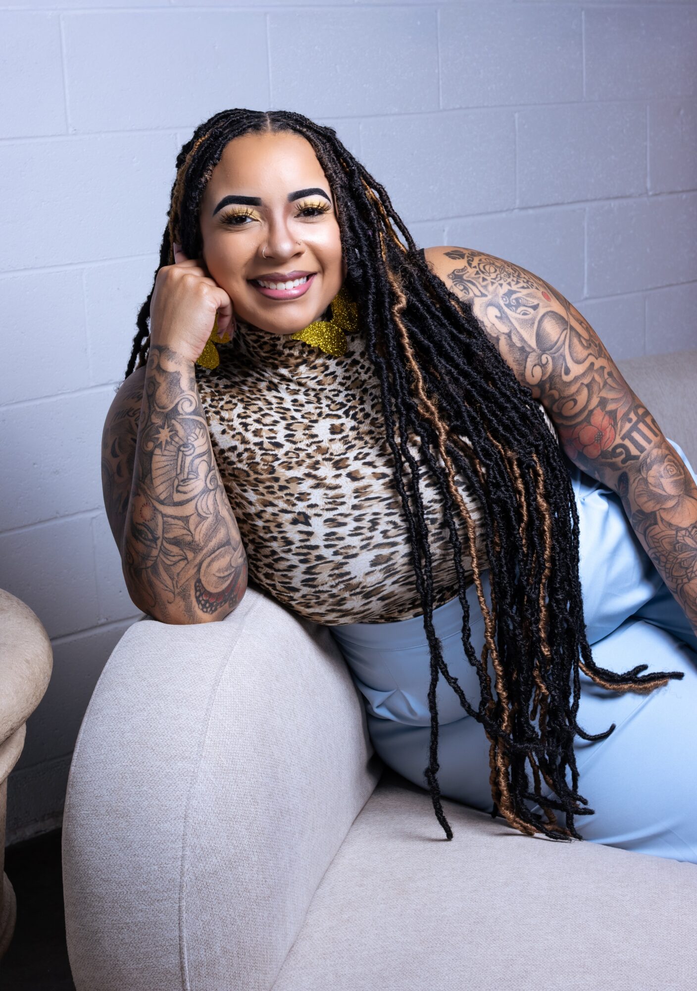 Smiling woman with long dreadlocks and tattoos, resting her head on her hand, sitting on a light-colored sofa against a gray wall.