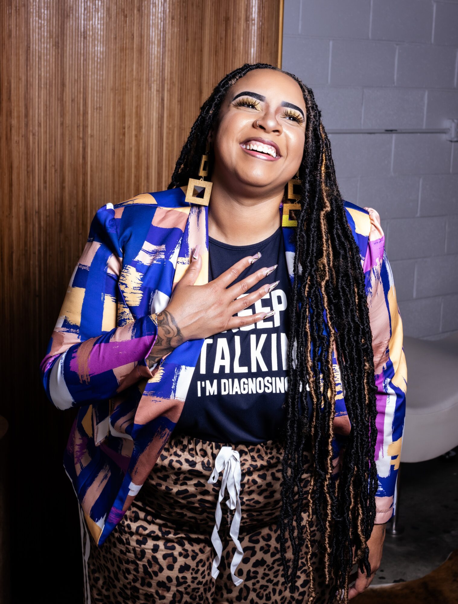 Woman with long dreadlocks smiling, wearing a colorful jacket and leopard print pants, standing indoors against a wooden wall and gray brick wall.
