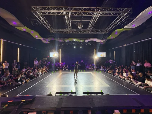 A dance or performance stage with a person in the center, surrounded by an audience in a dark room with colorful lighting.