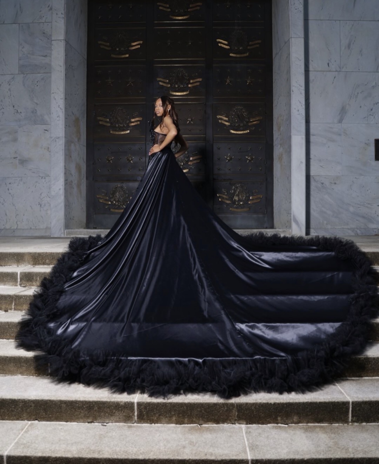 Woman in a black gown with a long train standing on steps in front of a large door.