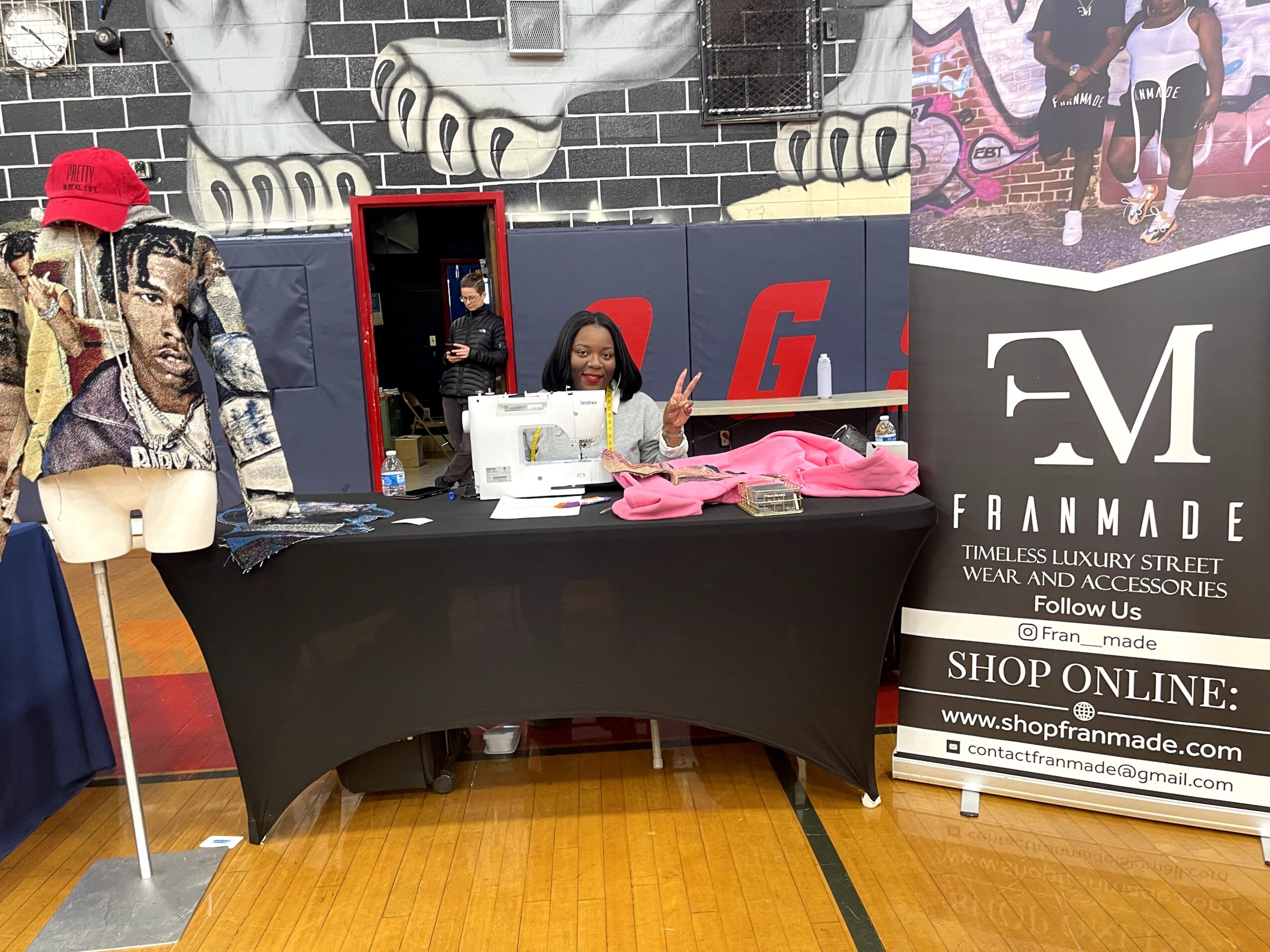Person sitting at a table with pink items, making a peace sign, in front of a mural and promotional banners.