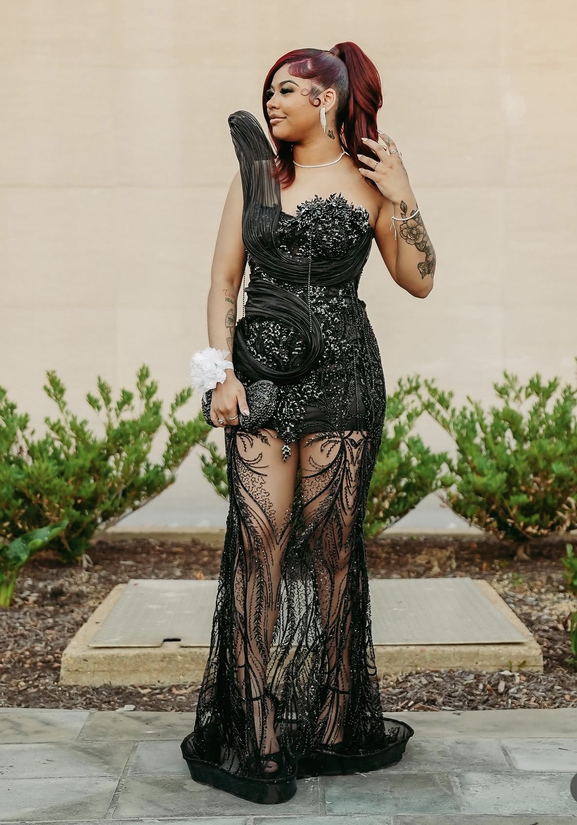 Woman in black lace dress with tattoos, standing outdoors with greenery and beige wall background.