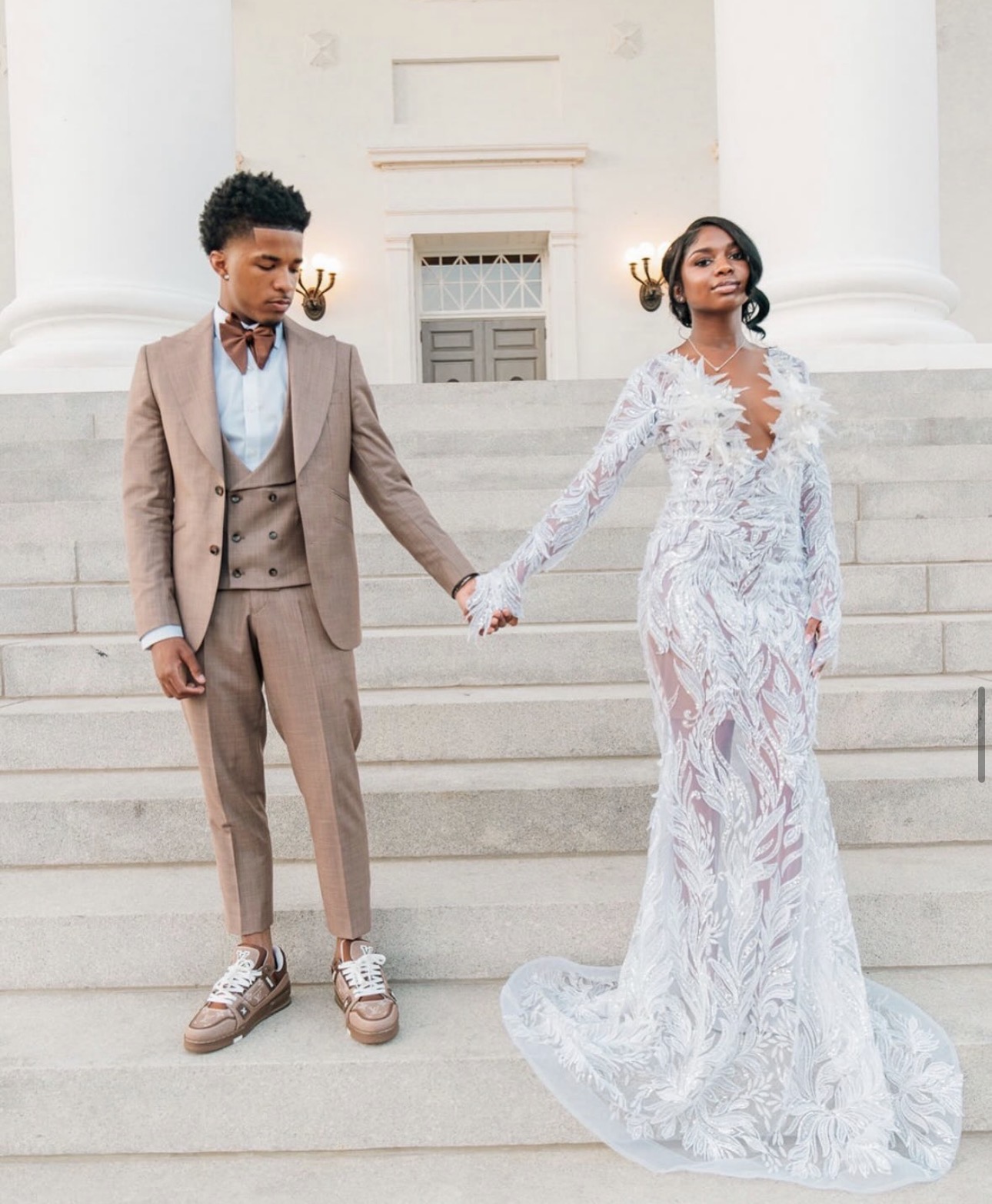 A couple holding hands on steps in front of a building with large columns, dressed in formal attire, facing each other.