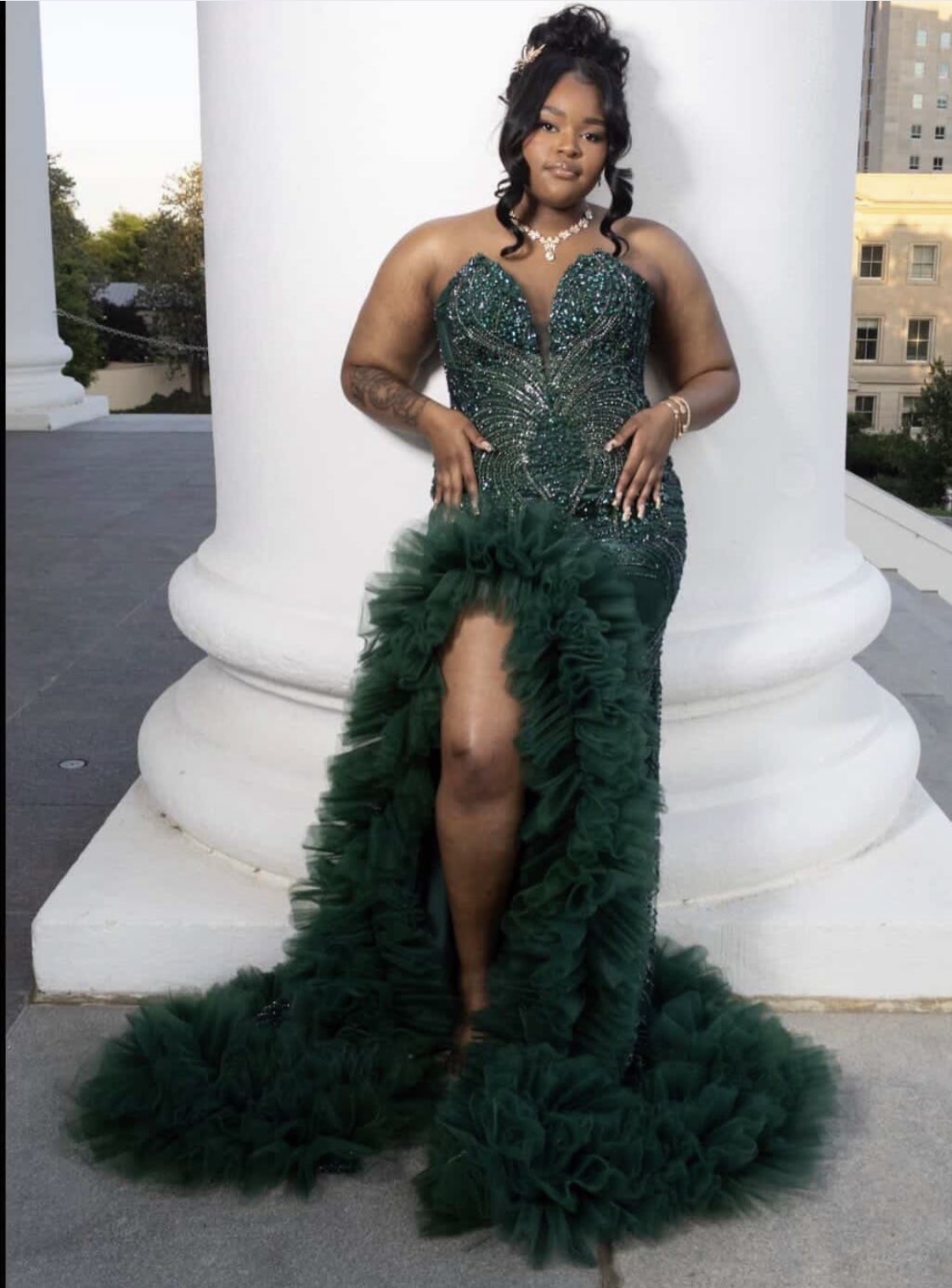 Woman in a green, beaded gown with a high slit, standing outdoors against a white column, city buildings in background.