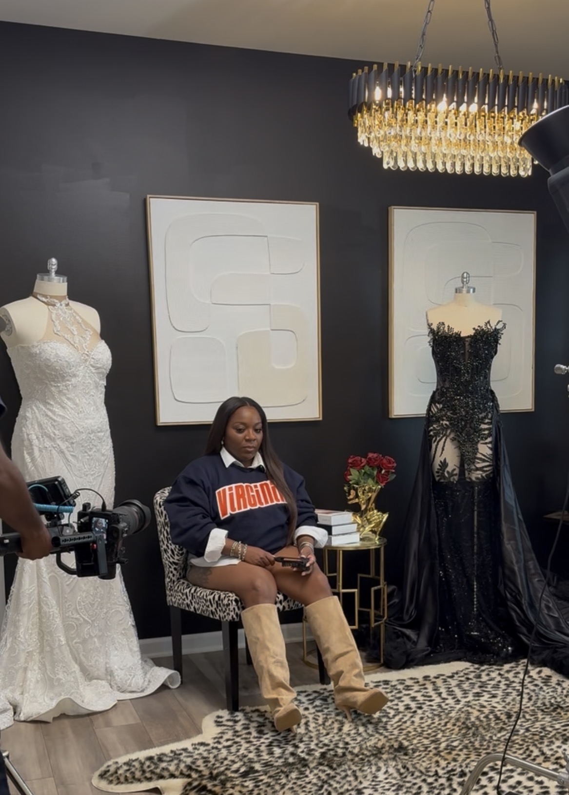 Woman sitting in a room with two mannequins wearing wedding dresses, dark wall, chandelier, and abstract art, holding a phone.