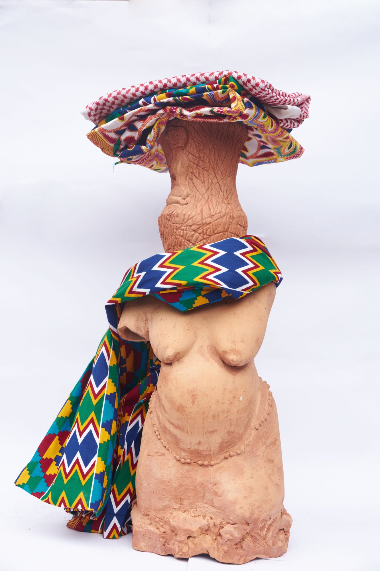 Sculpture of a person with a large head wearing colorful patterned fabric, standing on a textured base.