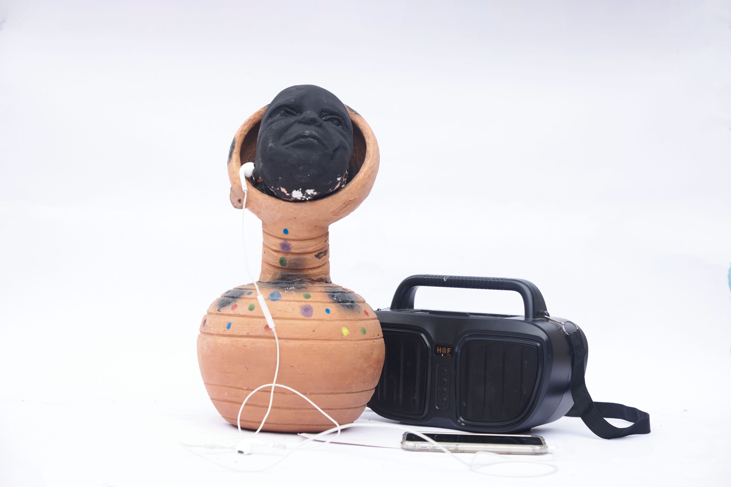 Clay sculpture with a human head and neck, headphones, and a black speaker, against a white background.