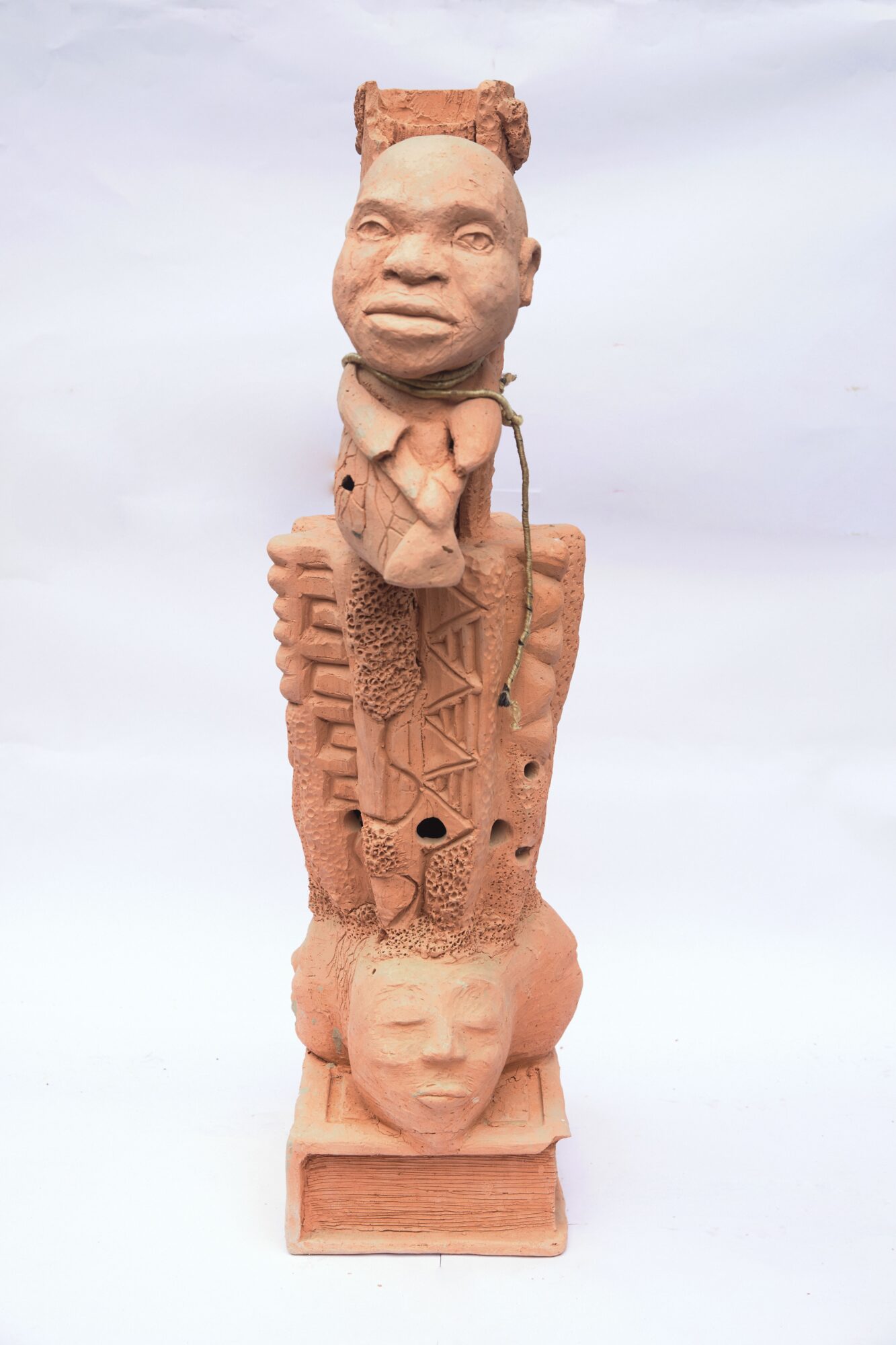 Tall sculpture with a human face on top, detailed carvings, and a smaller face at the base, made of reddish-brown material.