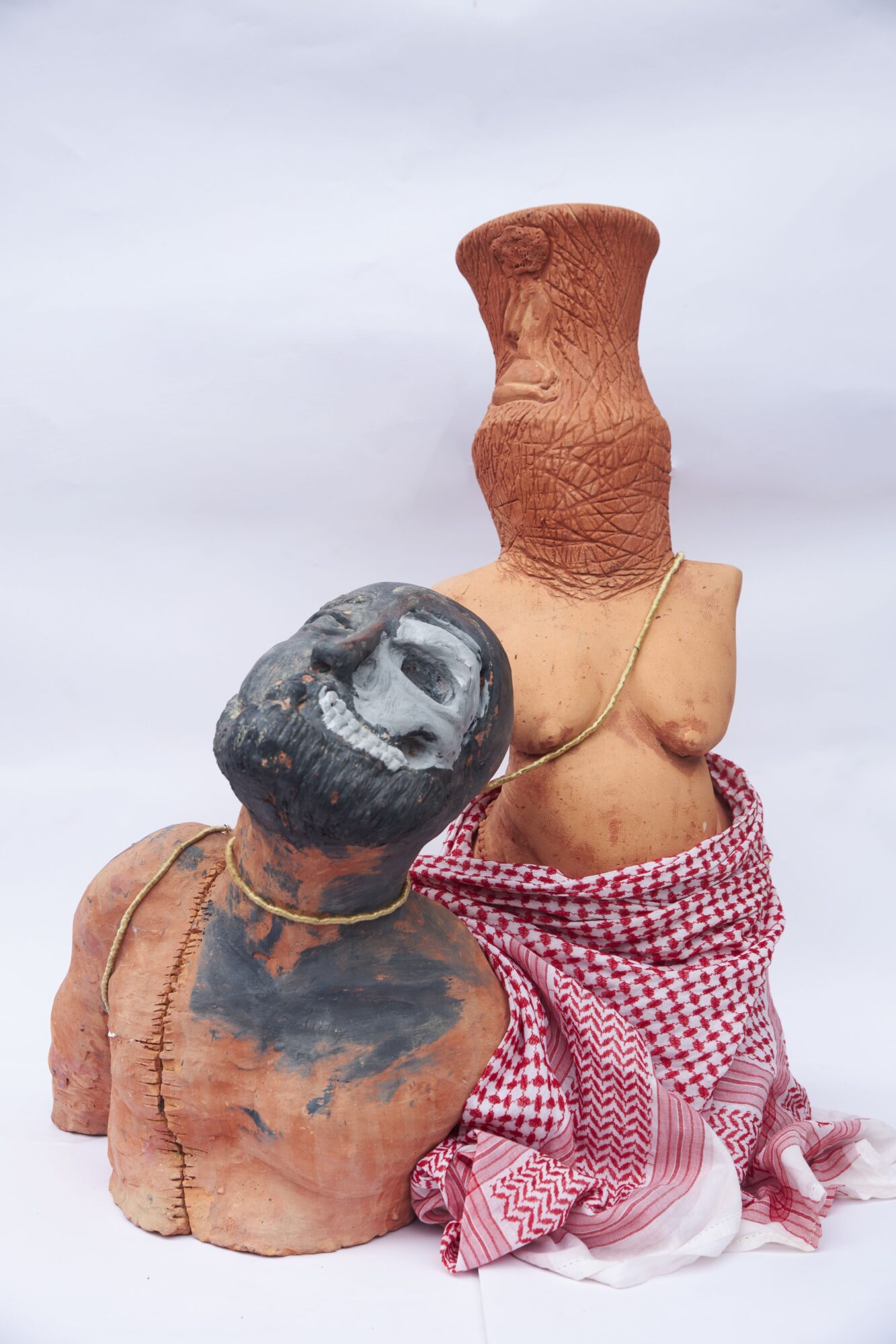 Two sculptures, one with a head and the other with a tall, textured torso, draped with a red and white cloth, against a cloudy sky.