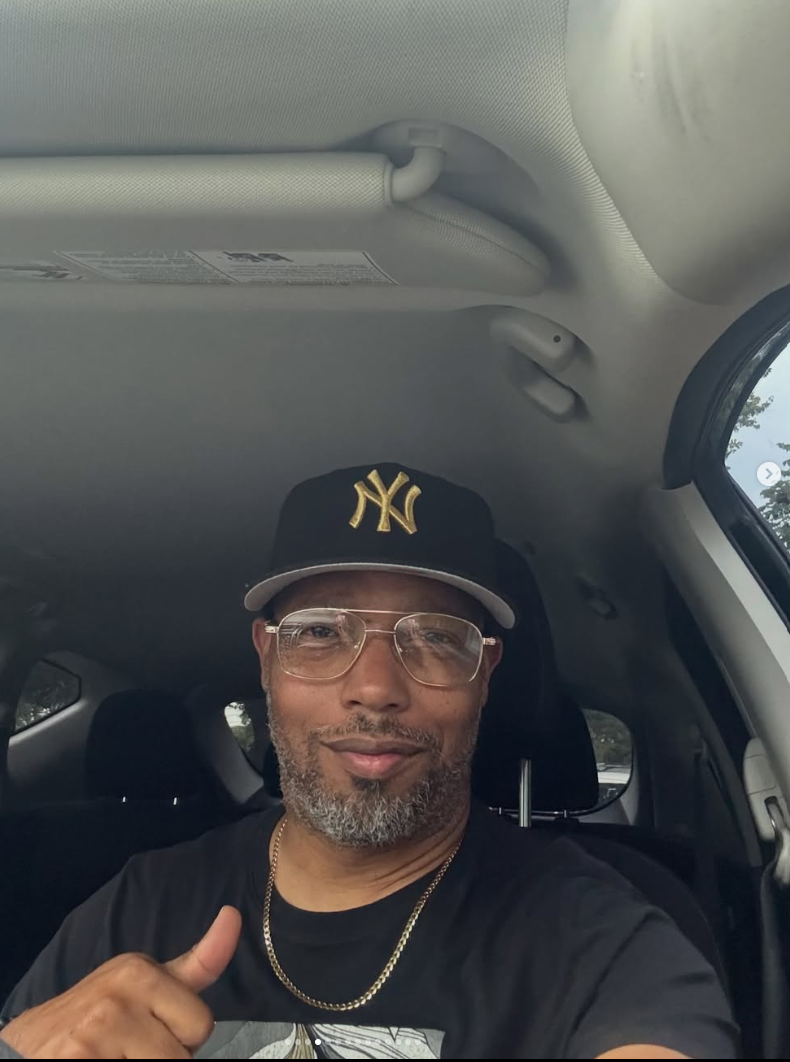 Man wearing glasses and a New York Yankees cap, sitting in a vehicle, smiling, with a beard and gold chain.