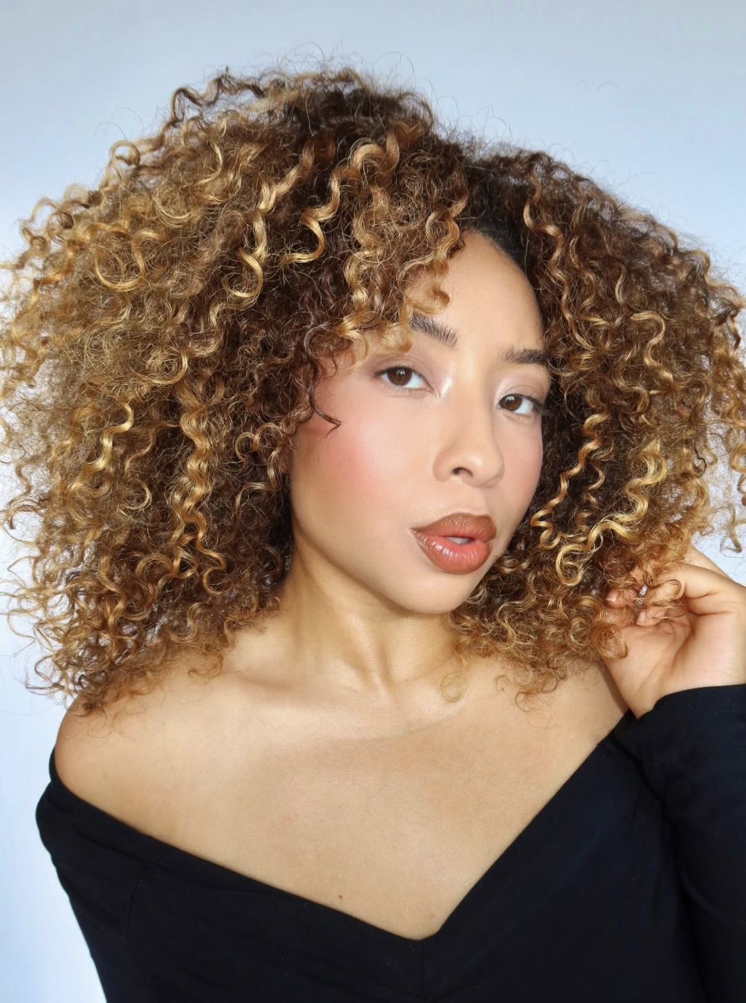 Woman with curly hair and off-shoulder black top, looking at camera, light blue background.