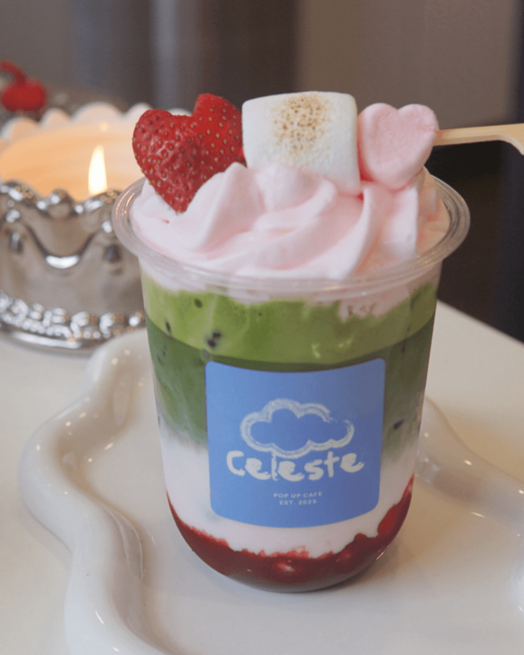 Layered dessert with pink whipped topping, strawberries, marshmallow, and green and white layers in a clear cup on a white tray.
