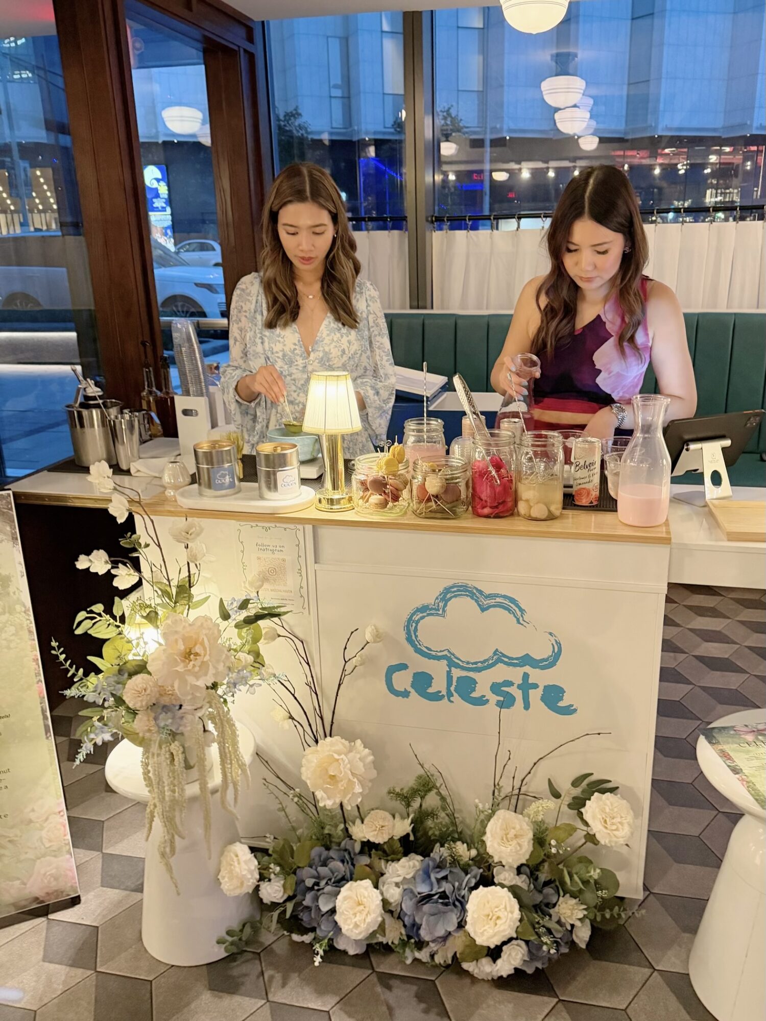 Two women stand behind a decorated counter with flowers and a sign that says 'Celeste' in a cafe setting.