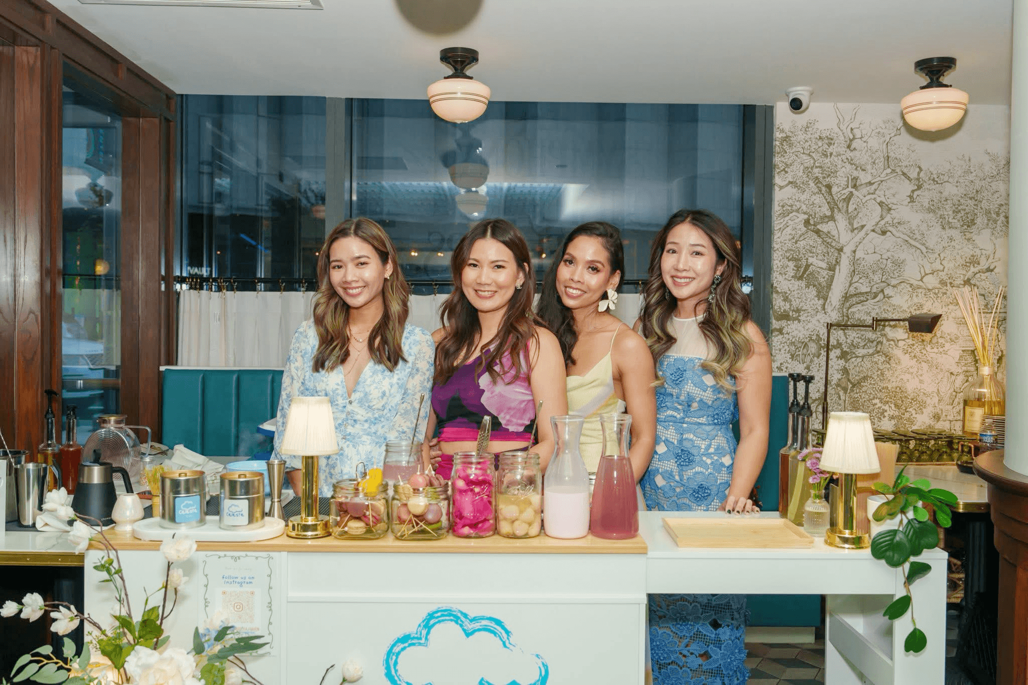 Four women stand behind a bar counter with drinks and decorations, smiling in a well-lit indoor setting.