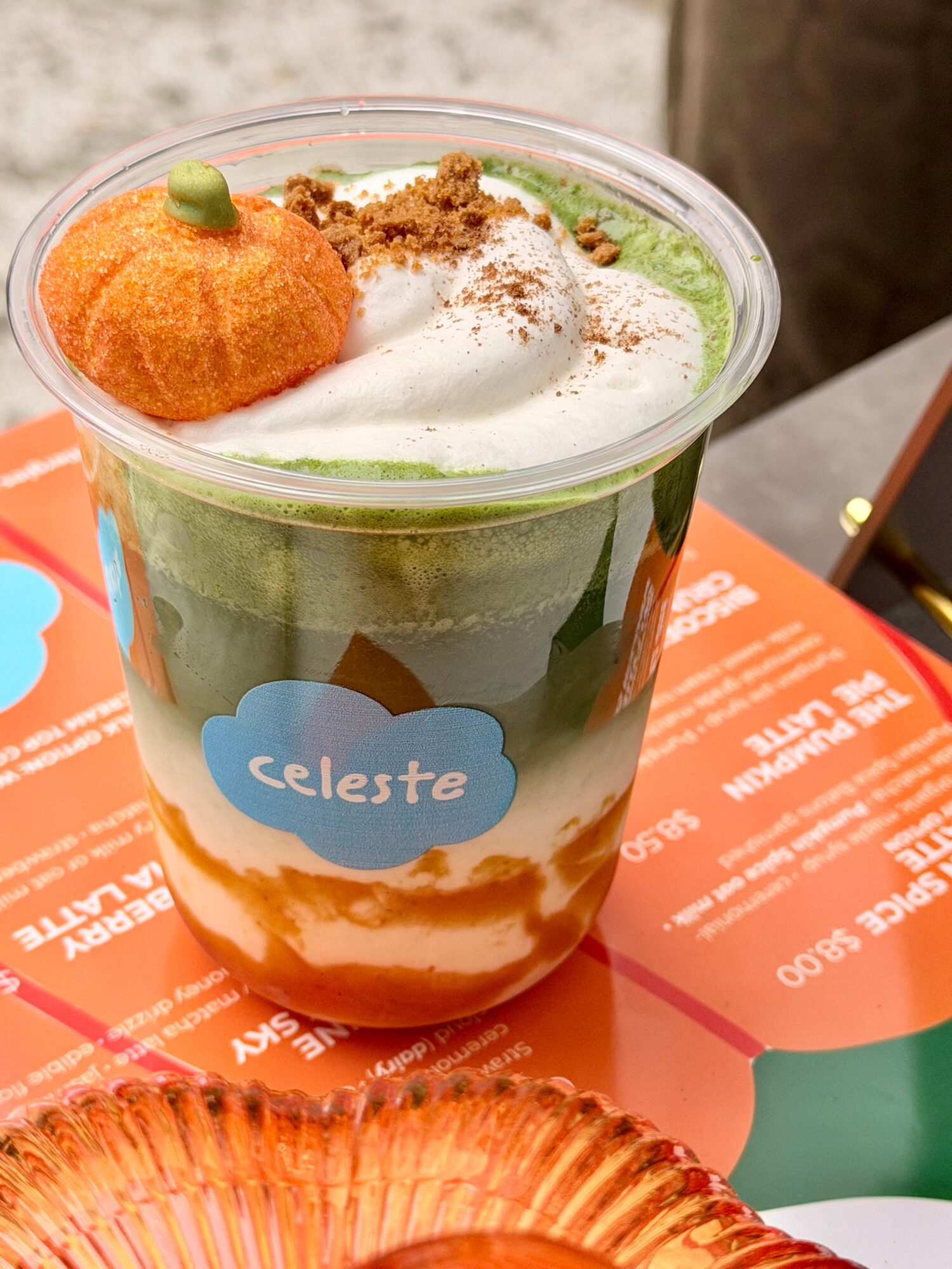 Layered dessert in a clear cup with green, white, and orange layers, topped with an orange decoration and sprinkled toppings.