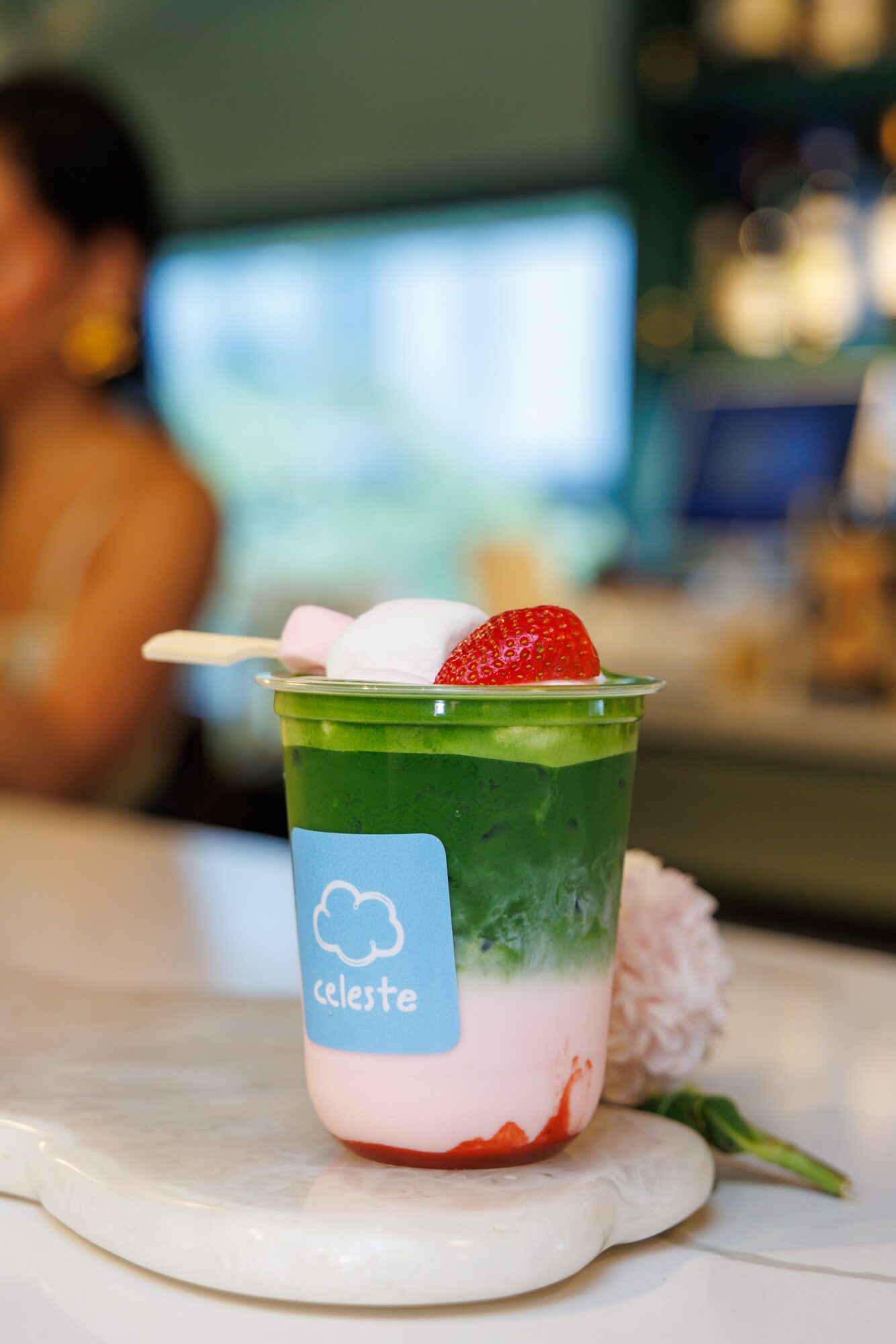 Layered green and pink drink with strawberries and marshmallows in a clear cup on a marble surface, blurred background.