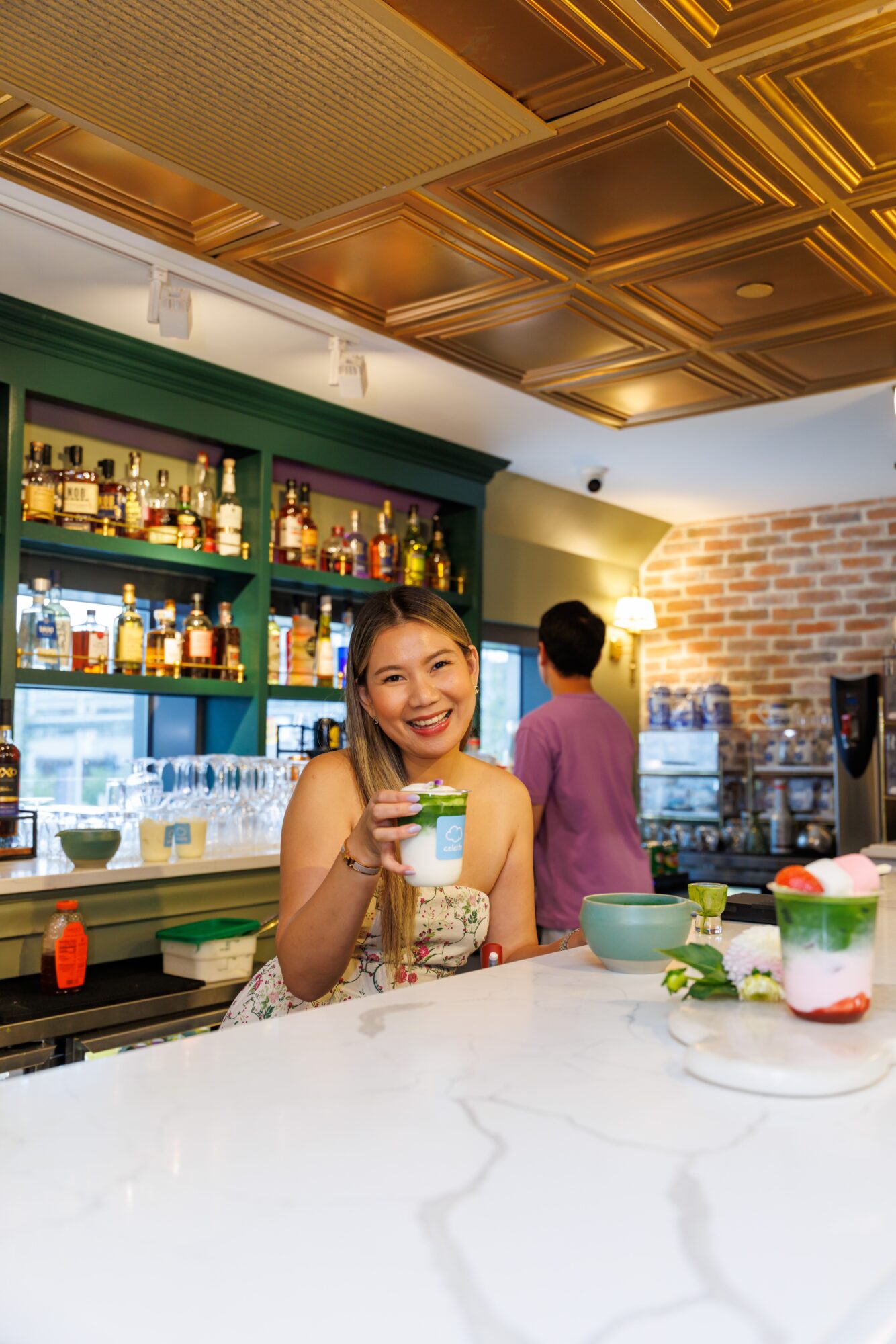Young woman smiling and holding a drink at a bar counter with colorful desserts and drinks, bar shelves in background.