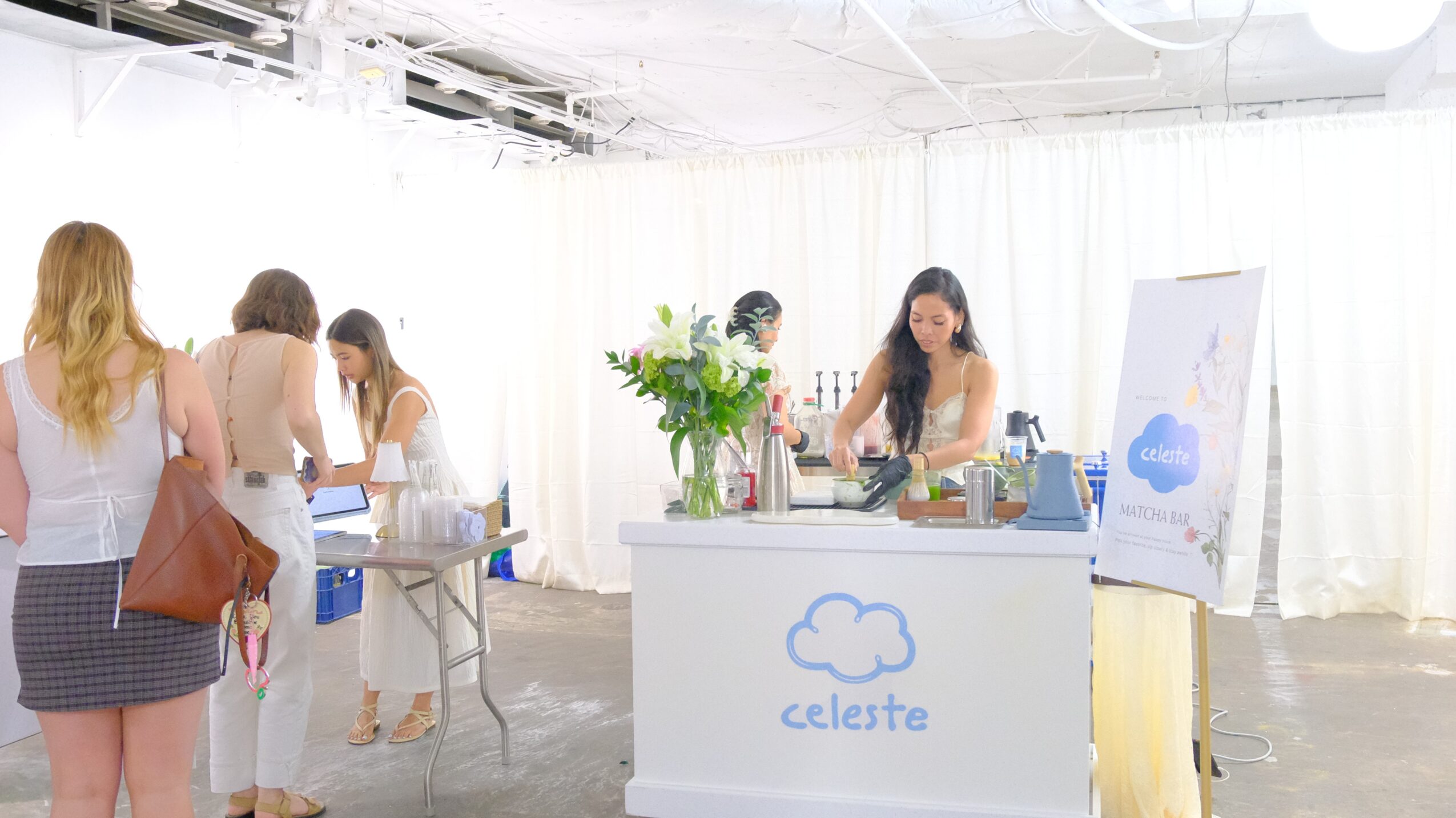 People gather around a booth with a cloud logo, a woman prepares drinks, and others wait in a bright, white space.