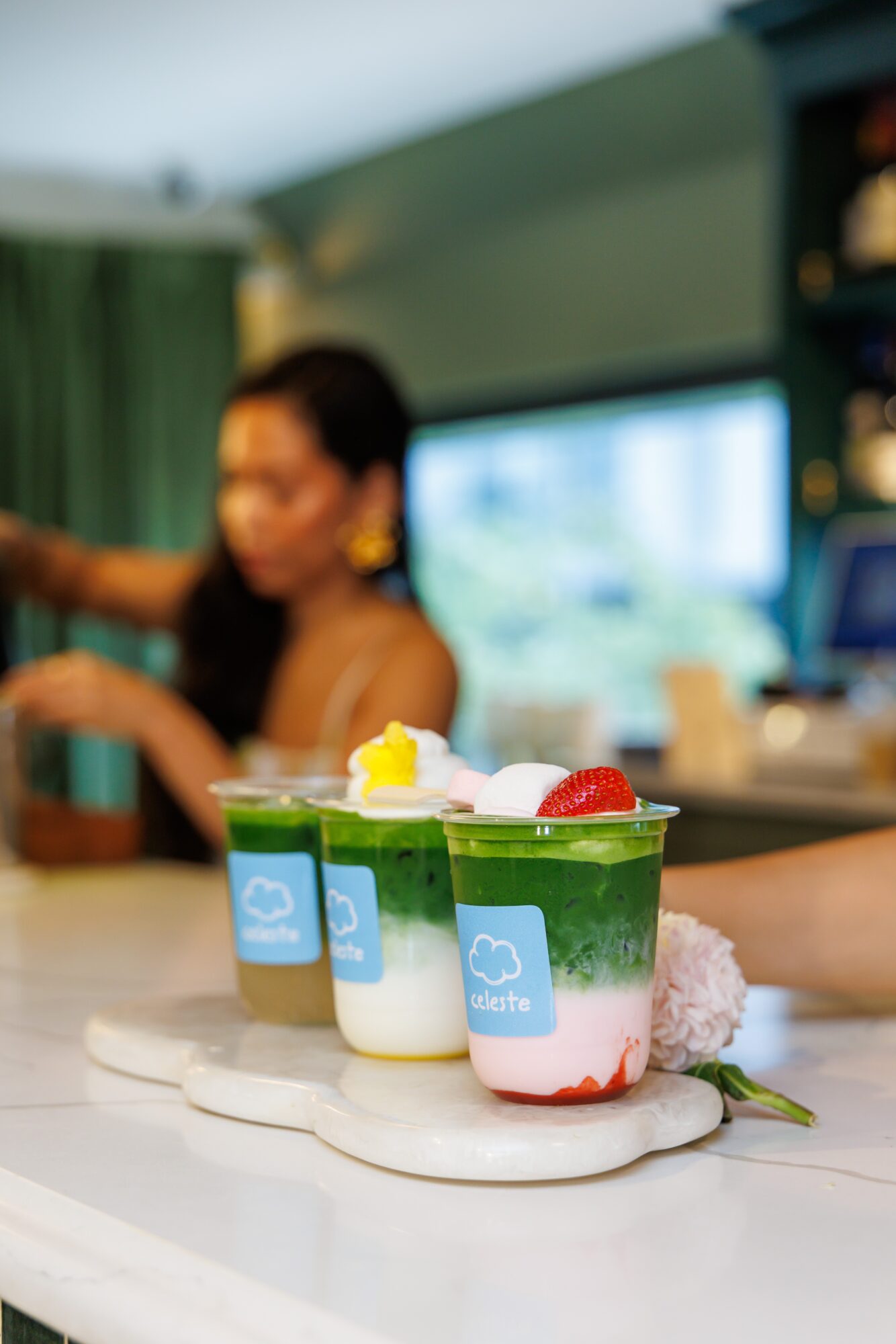Three colorful layered drinks with toppings on a white tray, blurred person in background, indoor setting.