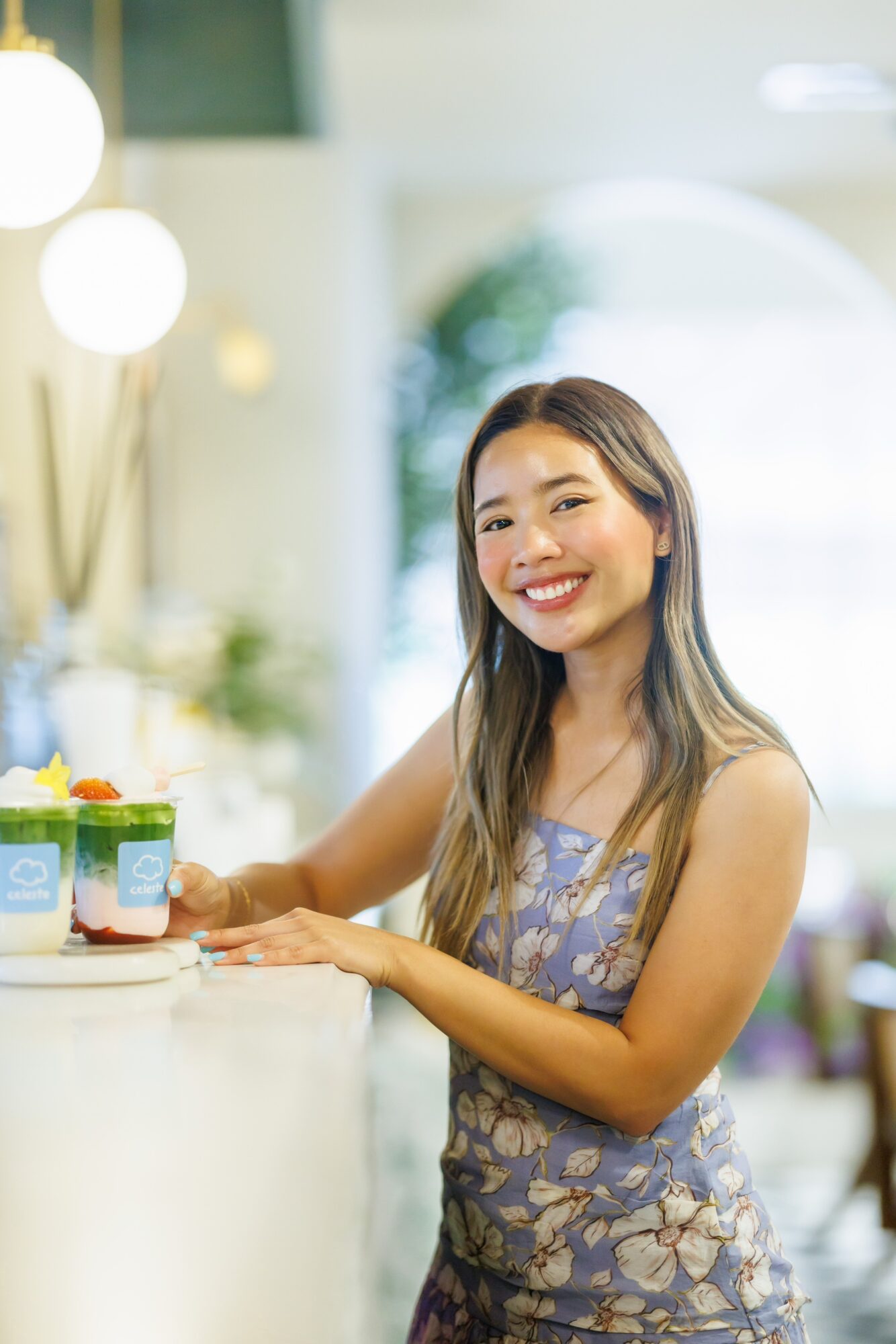 Smiling woman with long hair in a floral dress sitting at a table in a bright room.