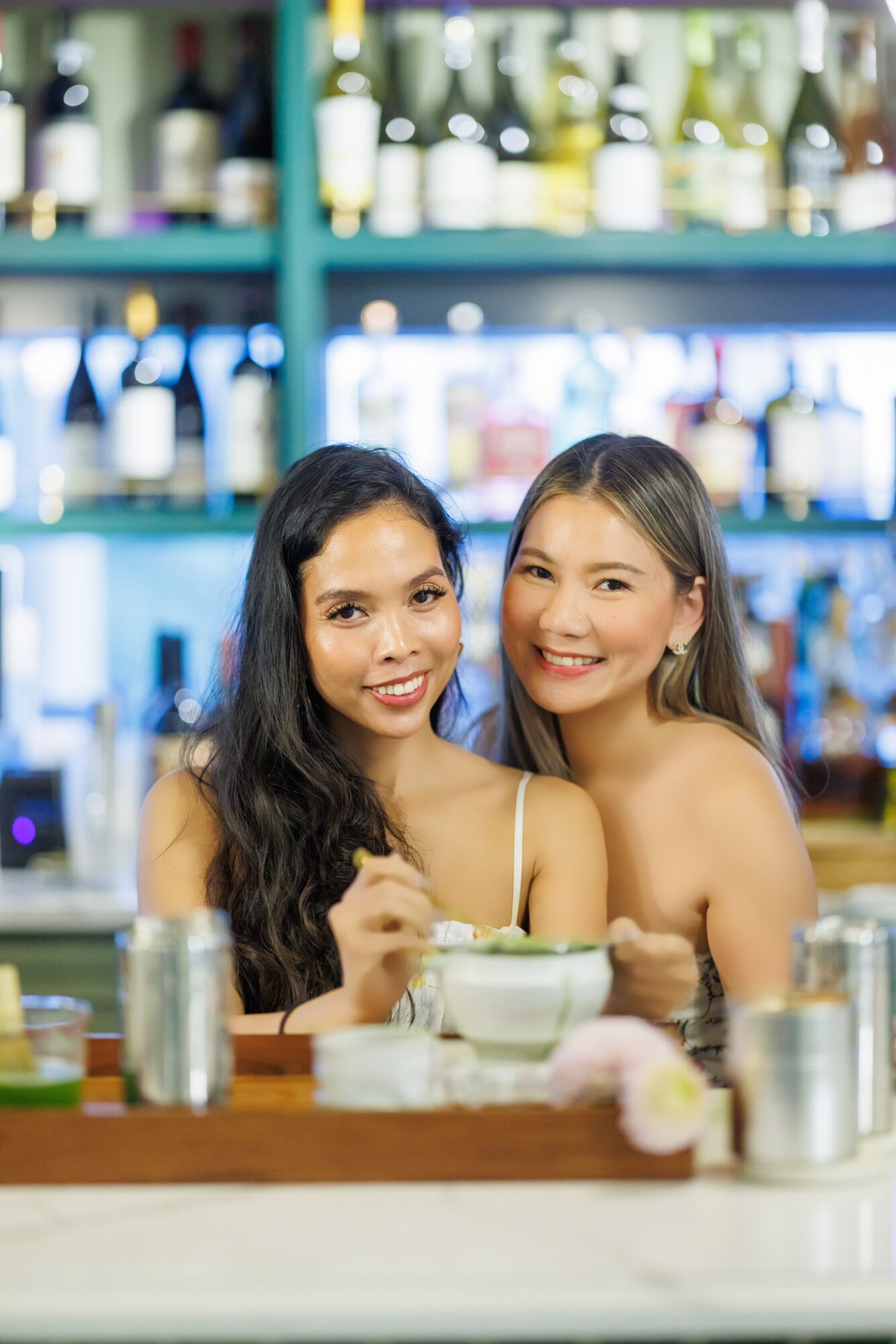 Two women smiling at a bar with bottles in the background, holding drinks, with bowls and glasses on the counter.