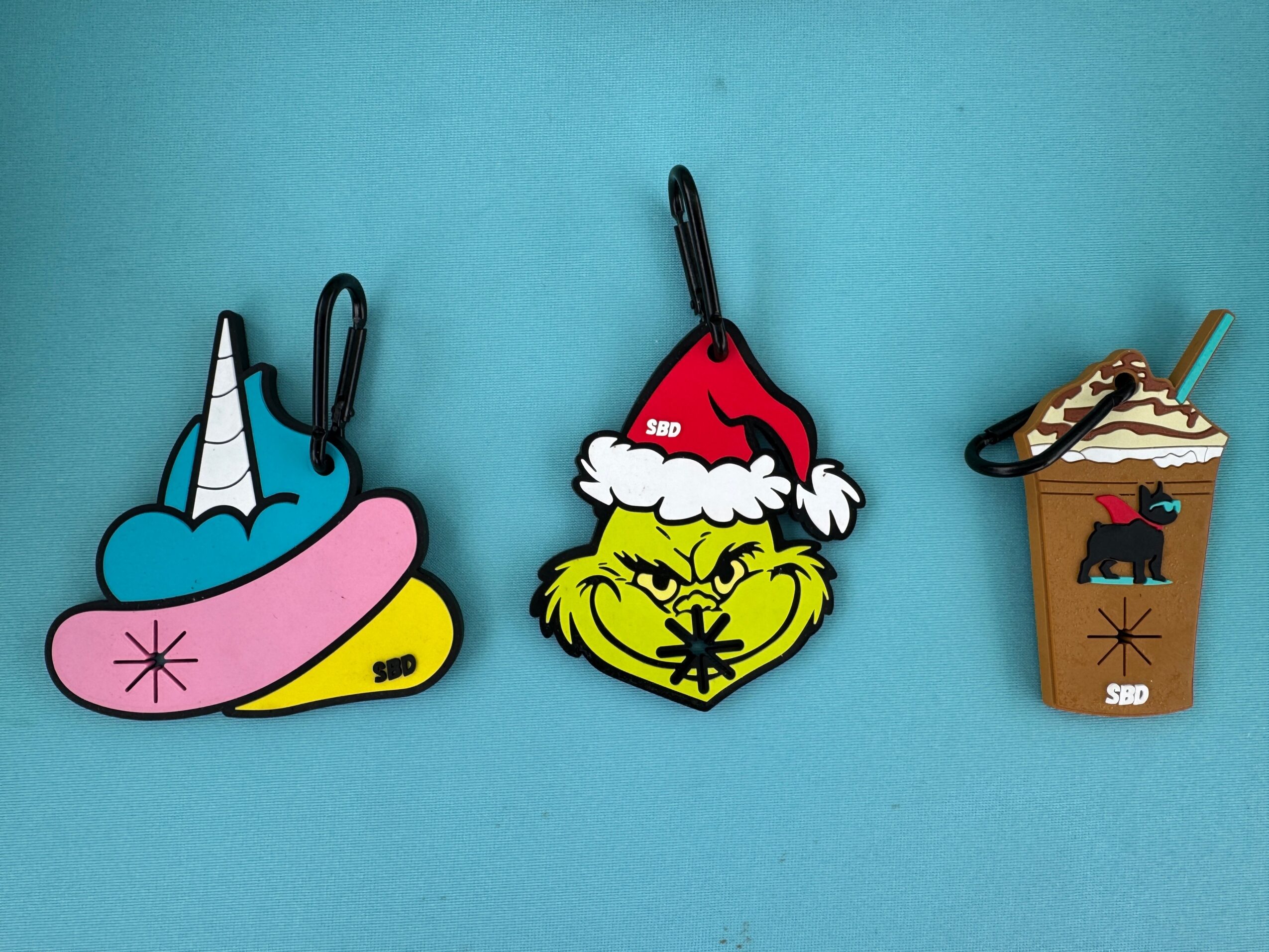Three holiday-themed keychains on a blue background: a unicorn with a pink mane, a Grinch with a Santa hat, and a coffee cup with a dog image.
