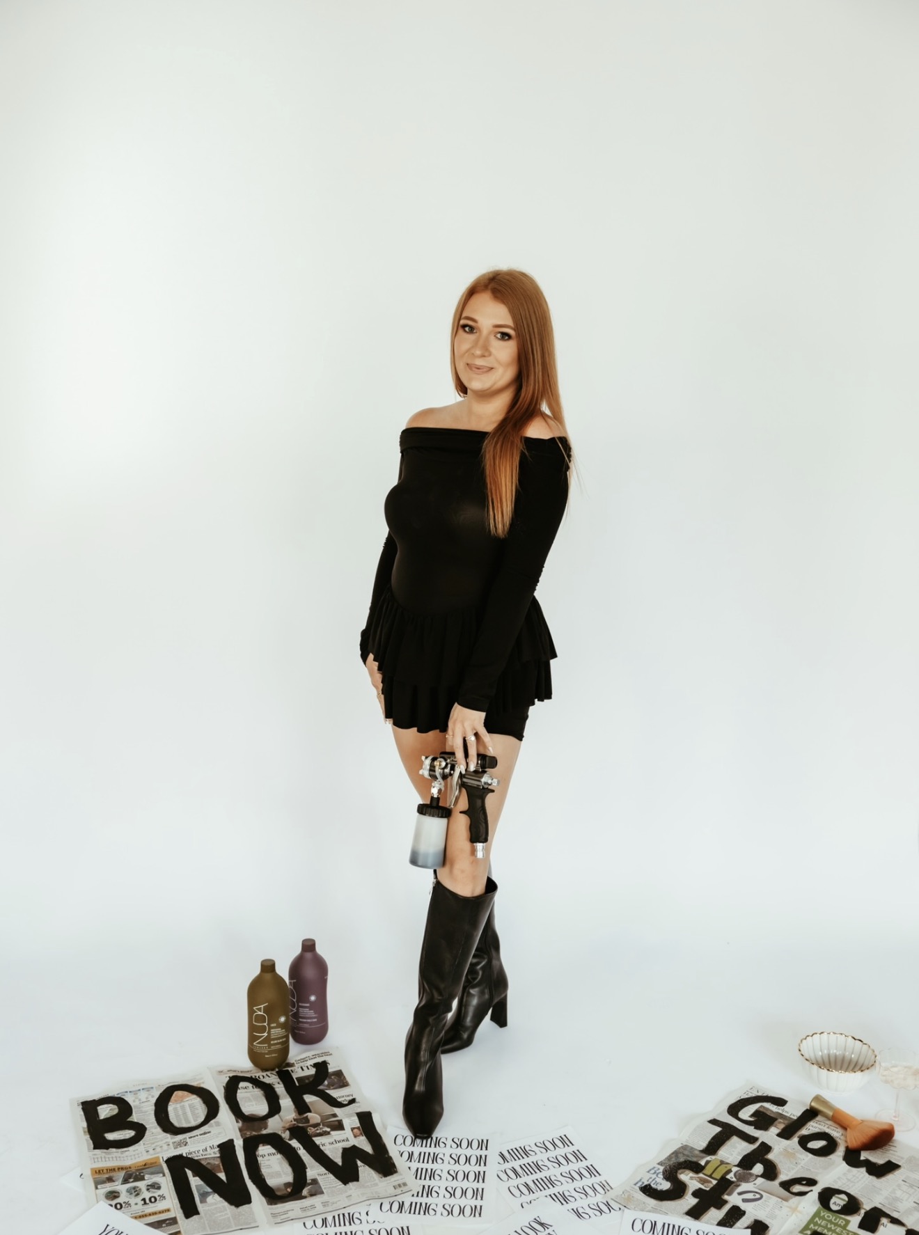 Woman in black dress standing with books and bottles on the floor, against a plain background.