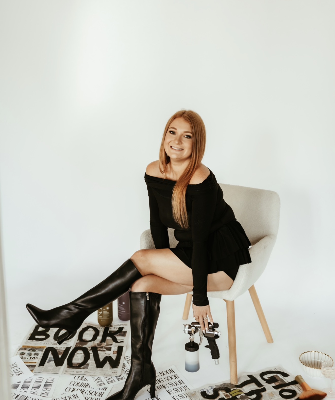 Woman with long hair sitting on a chair, holding a camera, smiling, in a minimalistic room with a white background.