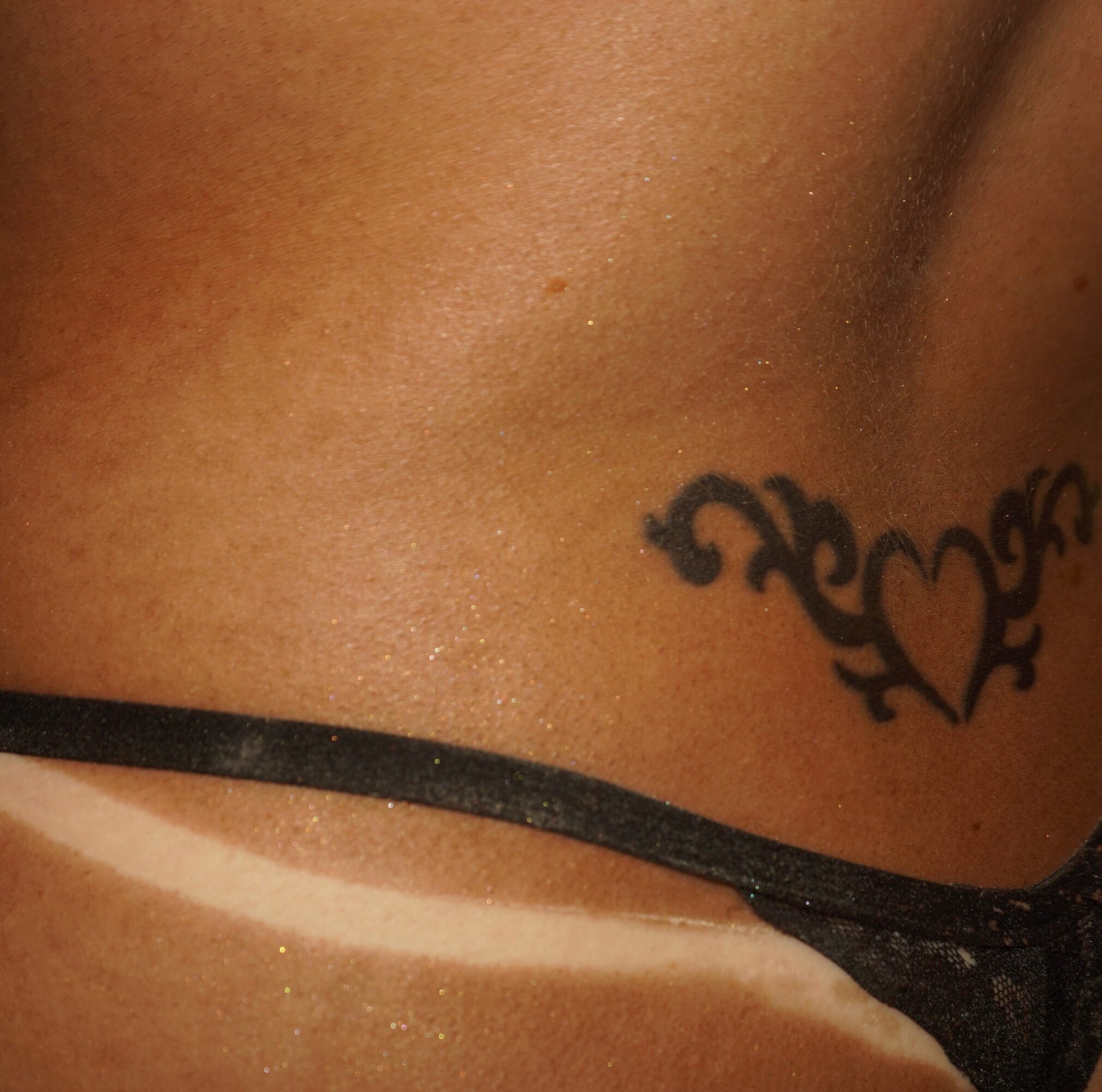 Close-up of skin with a black tattoo of a heart and decorative elements, and a black strap.