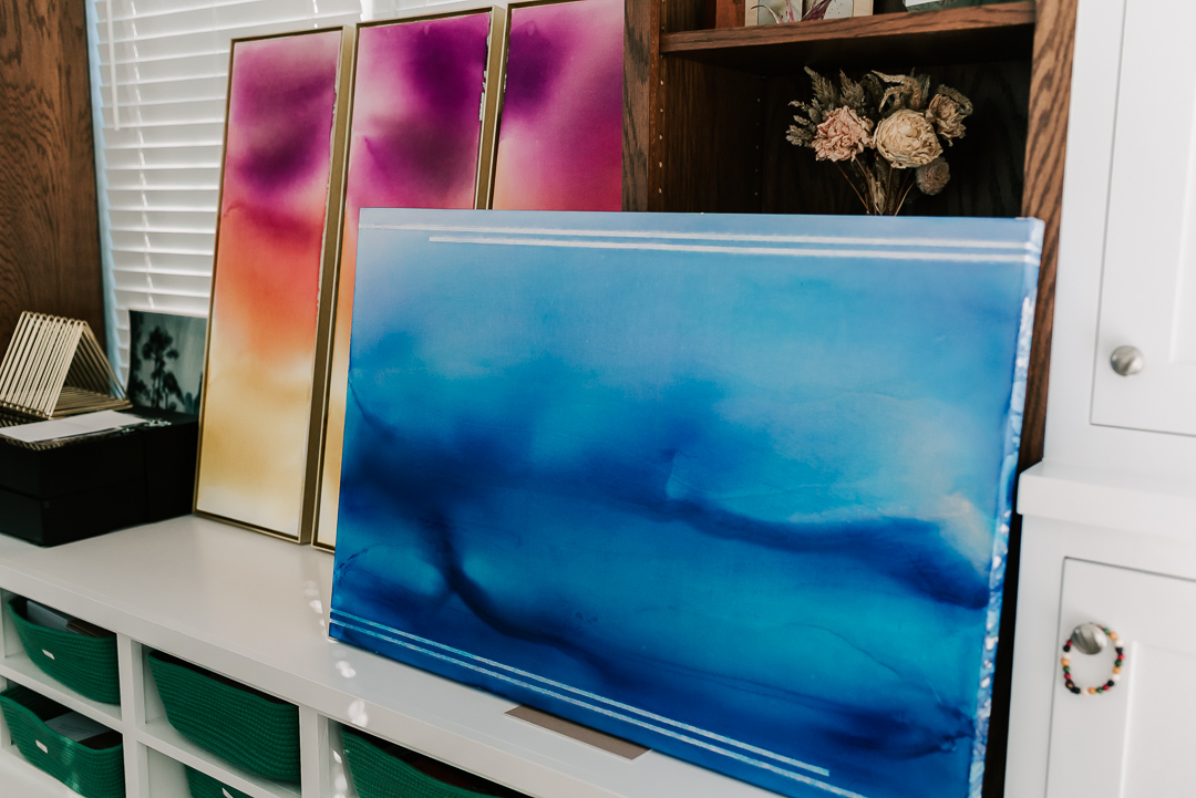 Three framed abstract paintings with pink and purple hues leaning against a wall, a blue landscape painting on a white shelf, and a wooden cabinet with flowers.
