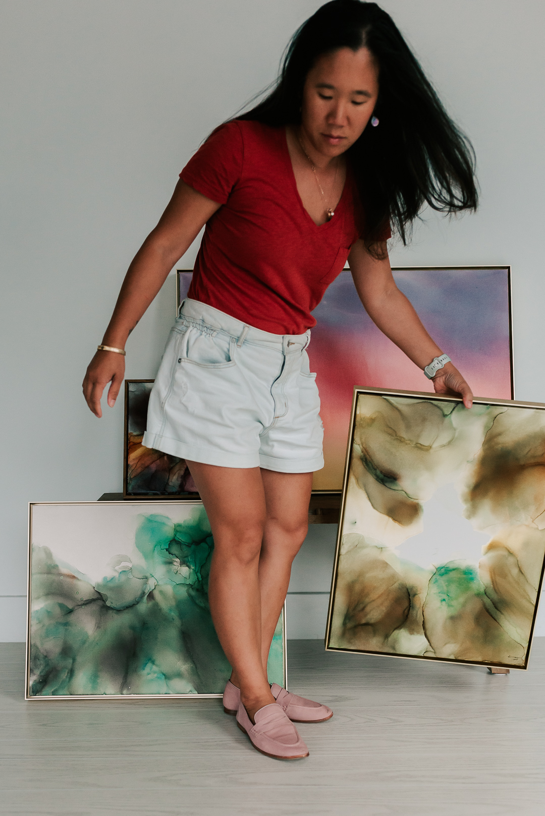 Woman in red shirt and white shorts looking down, standing among colorful abstract paintings, with hair blowing.