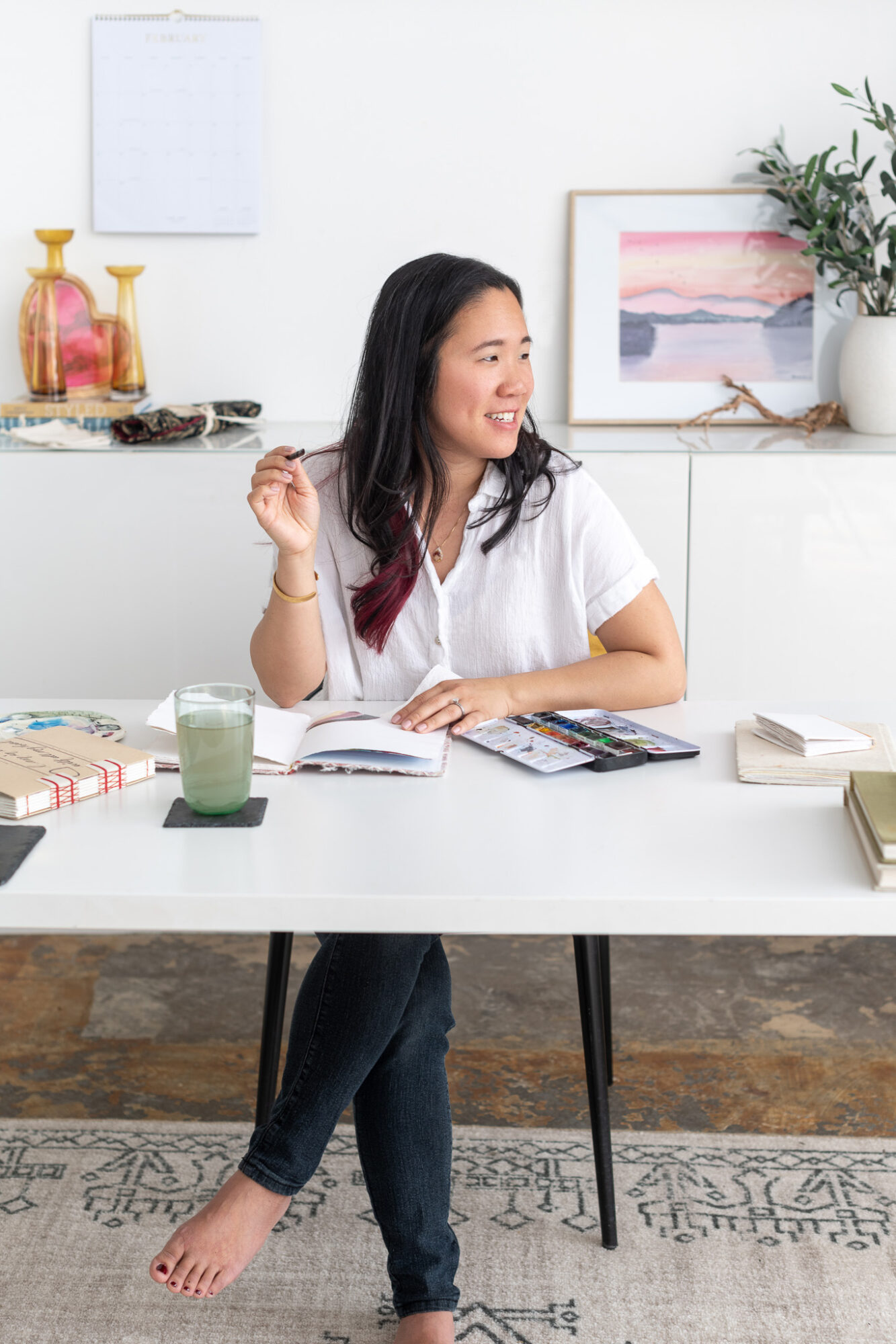 Woman sitting at a table with books, a glass of water, and a calculator, smiling and looking to the side.