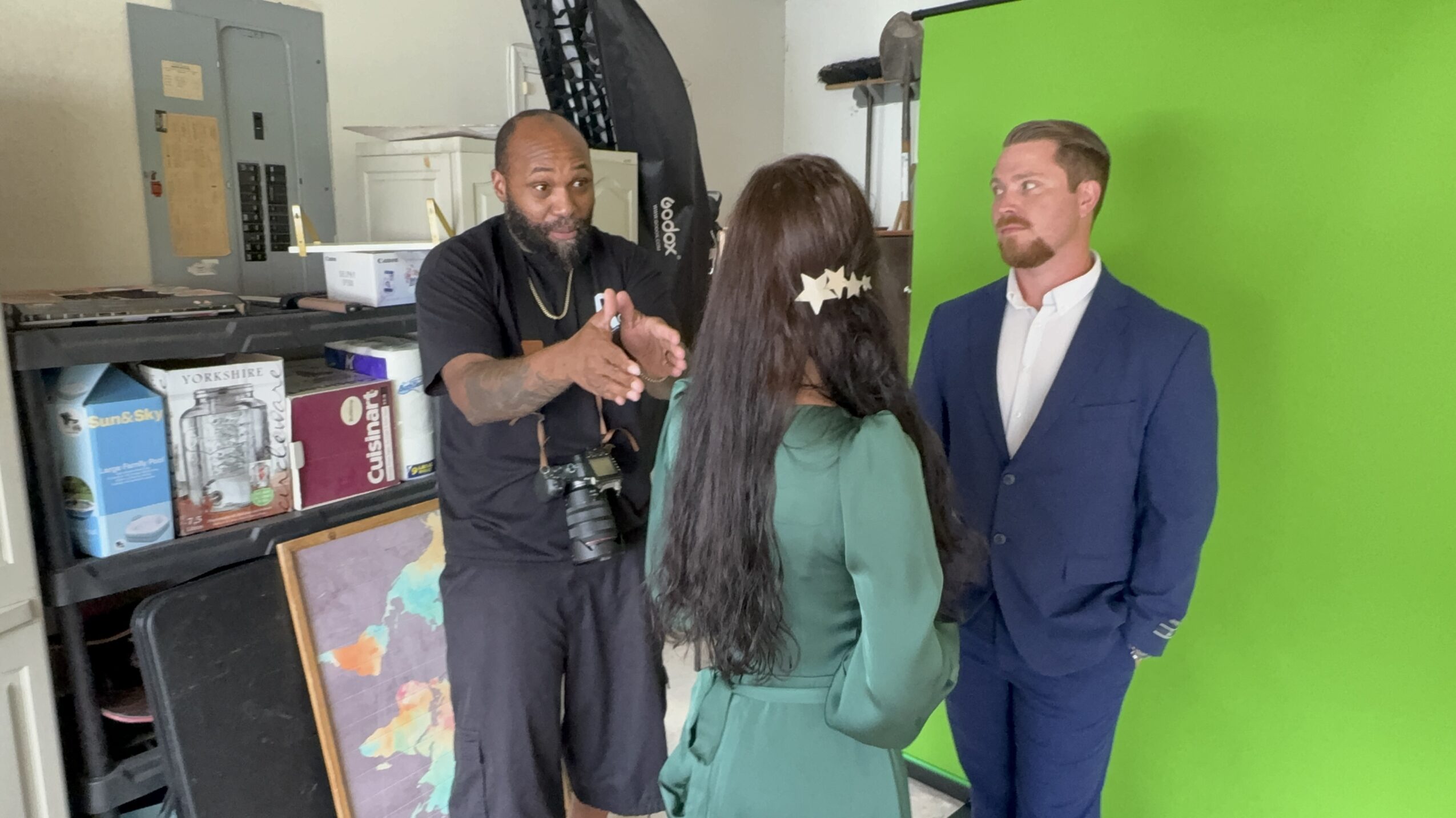 Three people in conversation in a studio with a green screen background, shelves, and equipment visible.