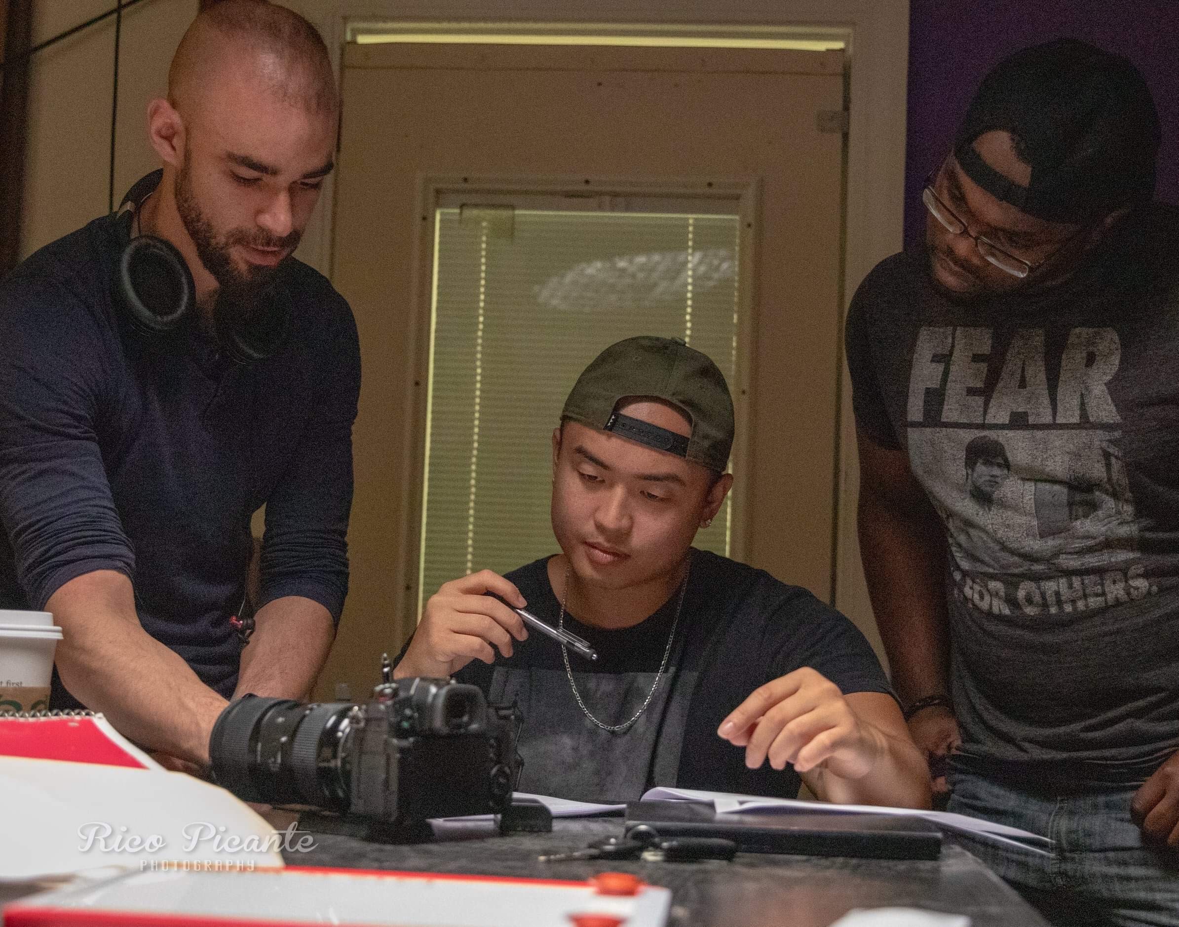 Three young men gathered around a table with cameras and notebooks, one wearing headphones, in a room with a window.