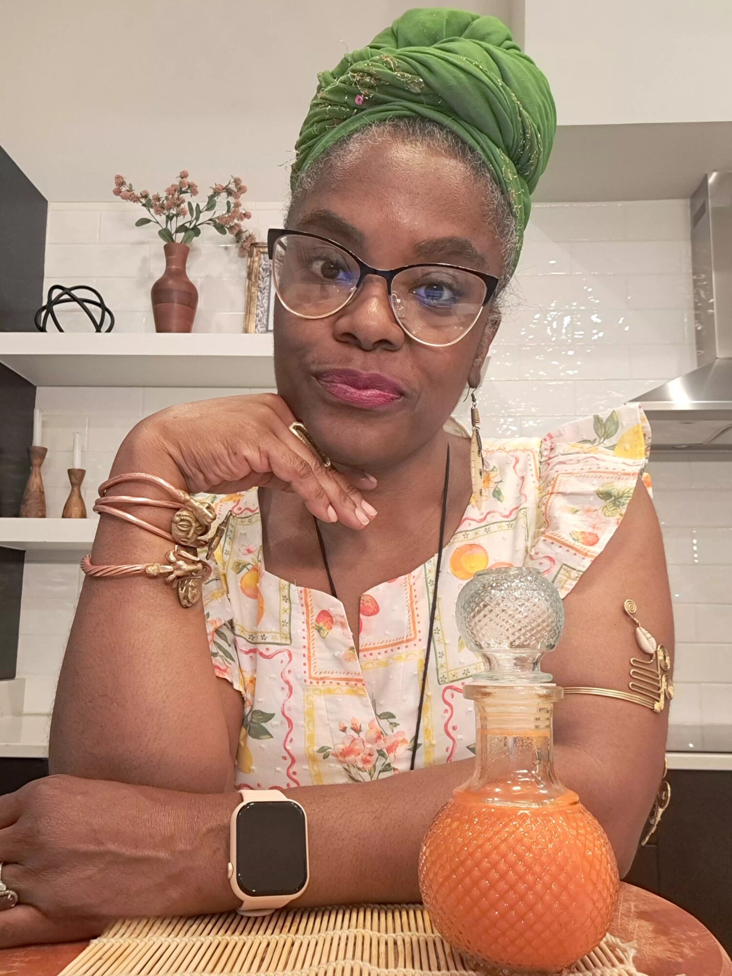 Woman wearing glasses and a green headwrap, sitting at a table with a decorative orange vase, in a room with shelves and plants.